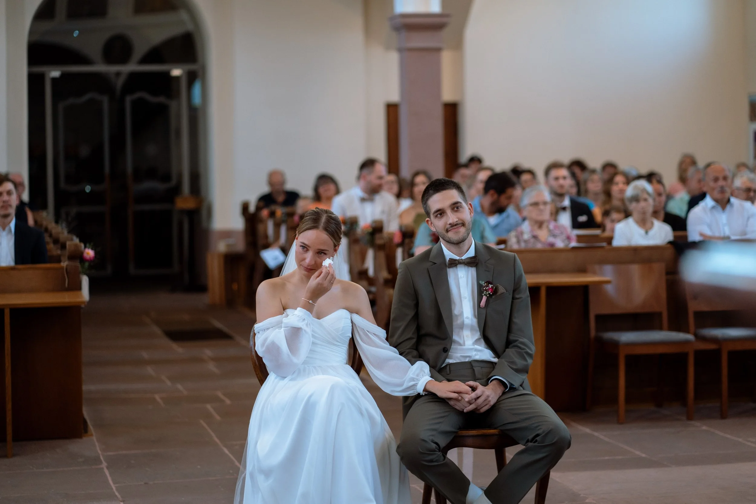 Bride and groom seated during a wedding ceremony inside a church, with guests watching in the background.