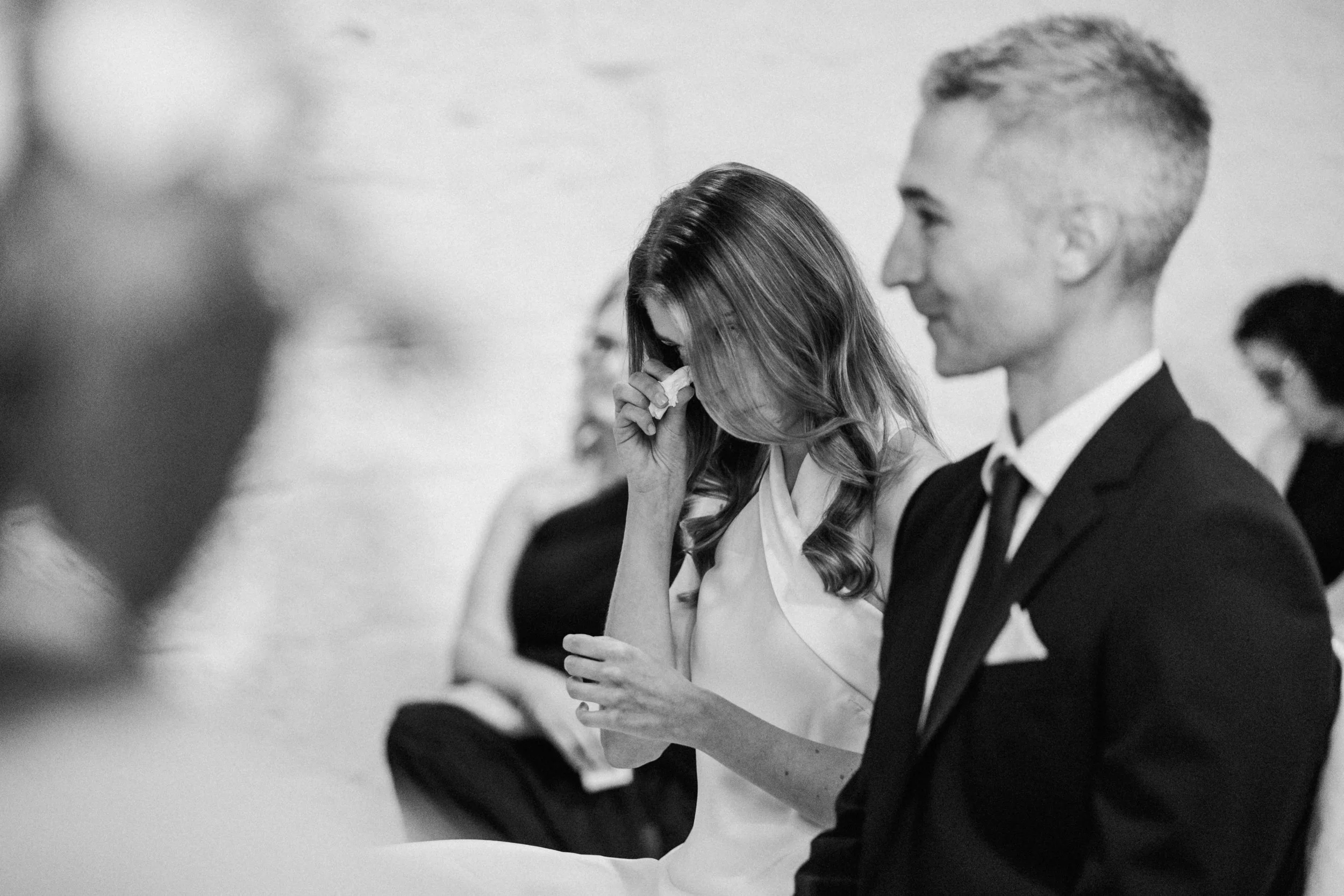 A black and white photo of a woman with long hair crying while holding a tissue to her face and a man with short hair smiling, both dressed in formal attire, at what appears to be a formal event or ceremony.