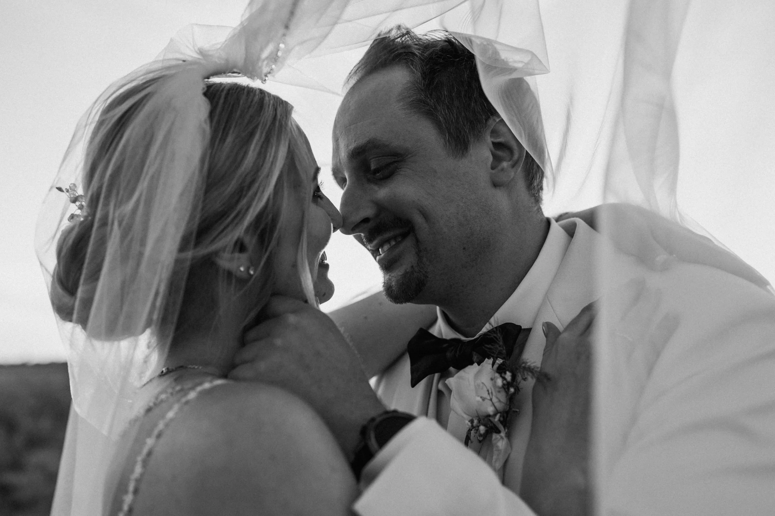 A black-and-white photograph of a bride and groom sharing a moment under a veil, with their foreheads touching, smiling, and embracing each other during their wedding.