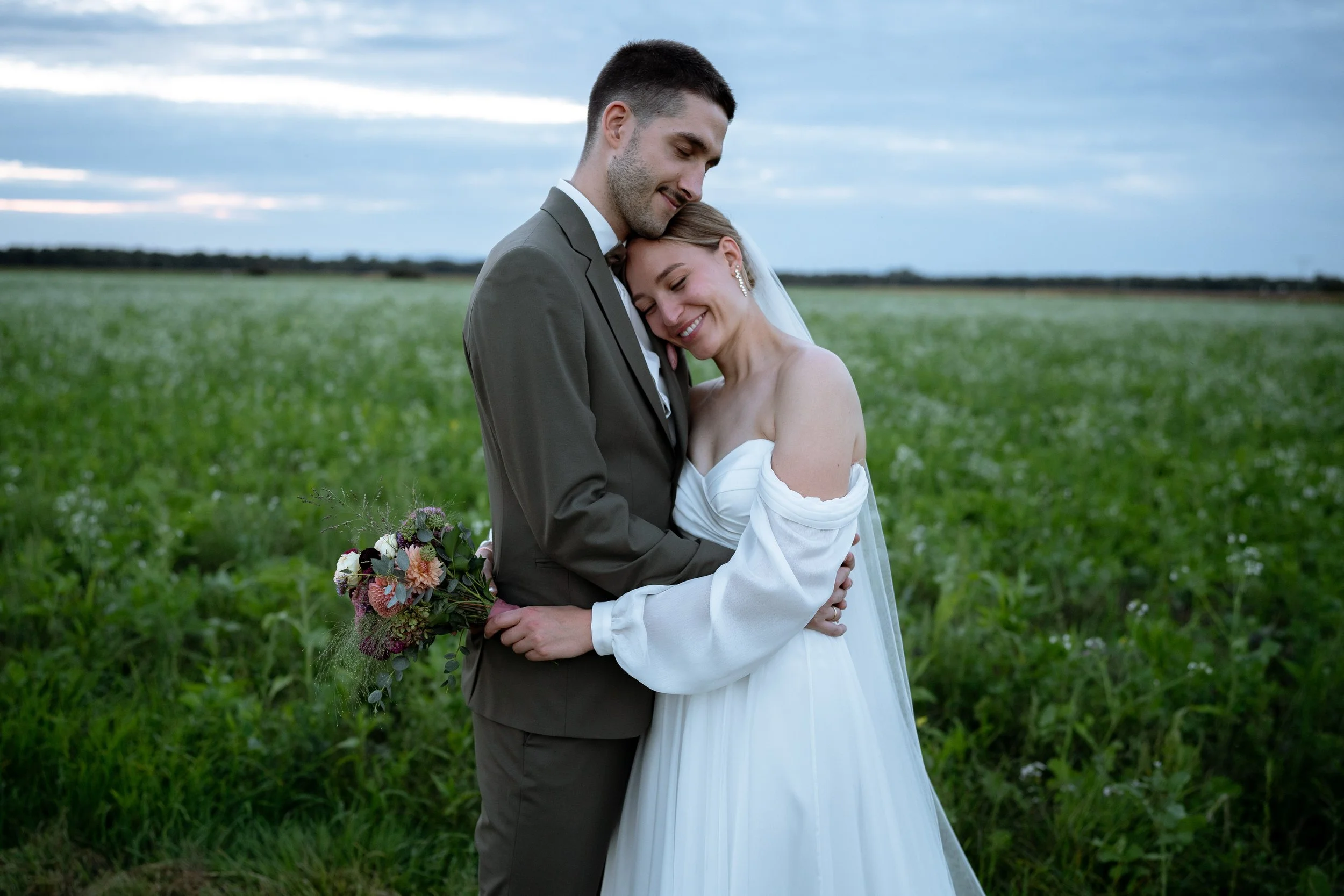 A bride and groom embrace in a field on their wedding day, with the bride holding a bouquet of flowers and both smiling with closed eyes.