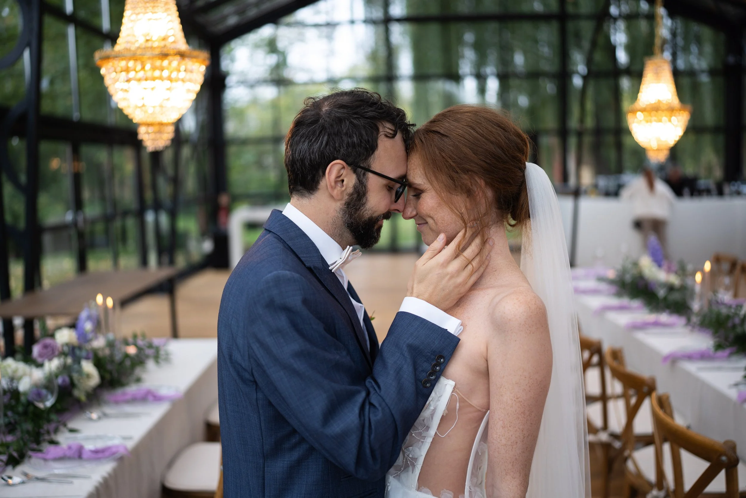 A bride and groom are touching foreheads and smiling at each other during their wedding ceremony in a decorated outdoor venue with chandeliers and floral arrangements.