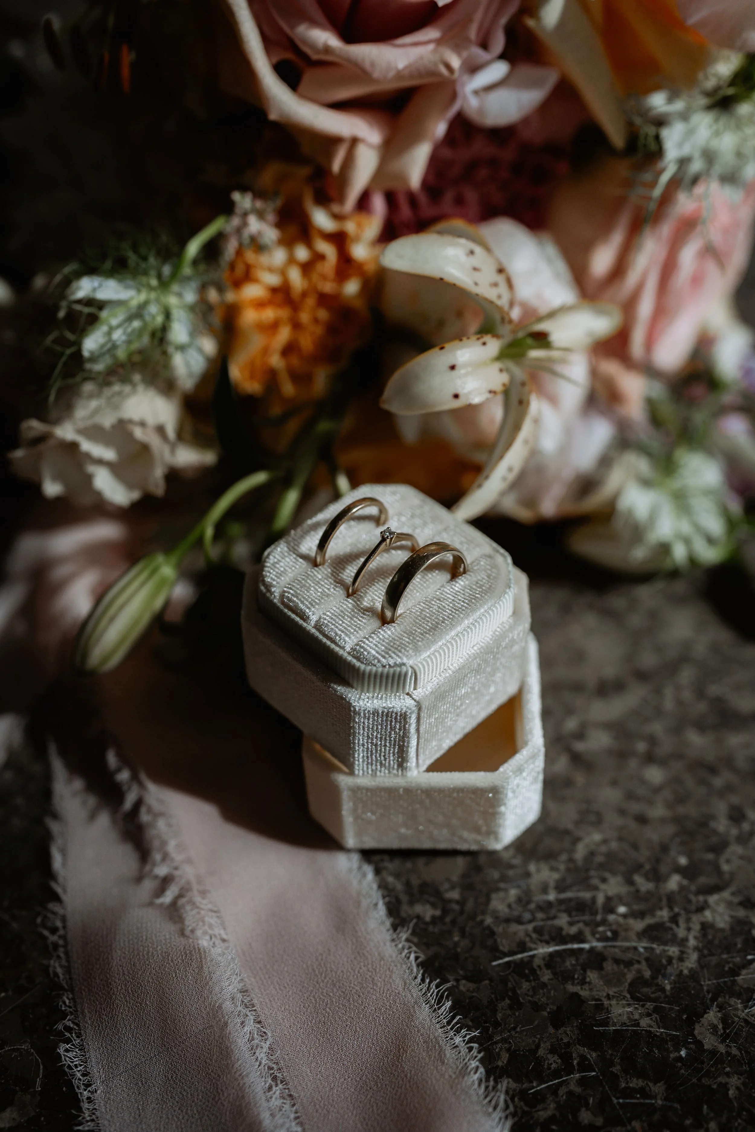 Close-up of two wedding bands on a white jewelry box, surrounded by white lilies, orange flowers, and greenery, with a pink fabric and a frayed light pink cloth underneath on a dark textured surface.