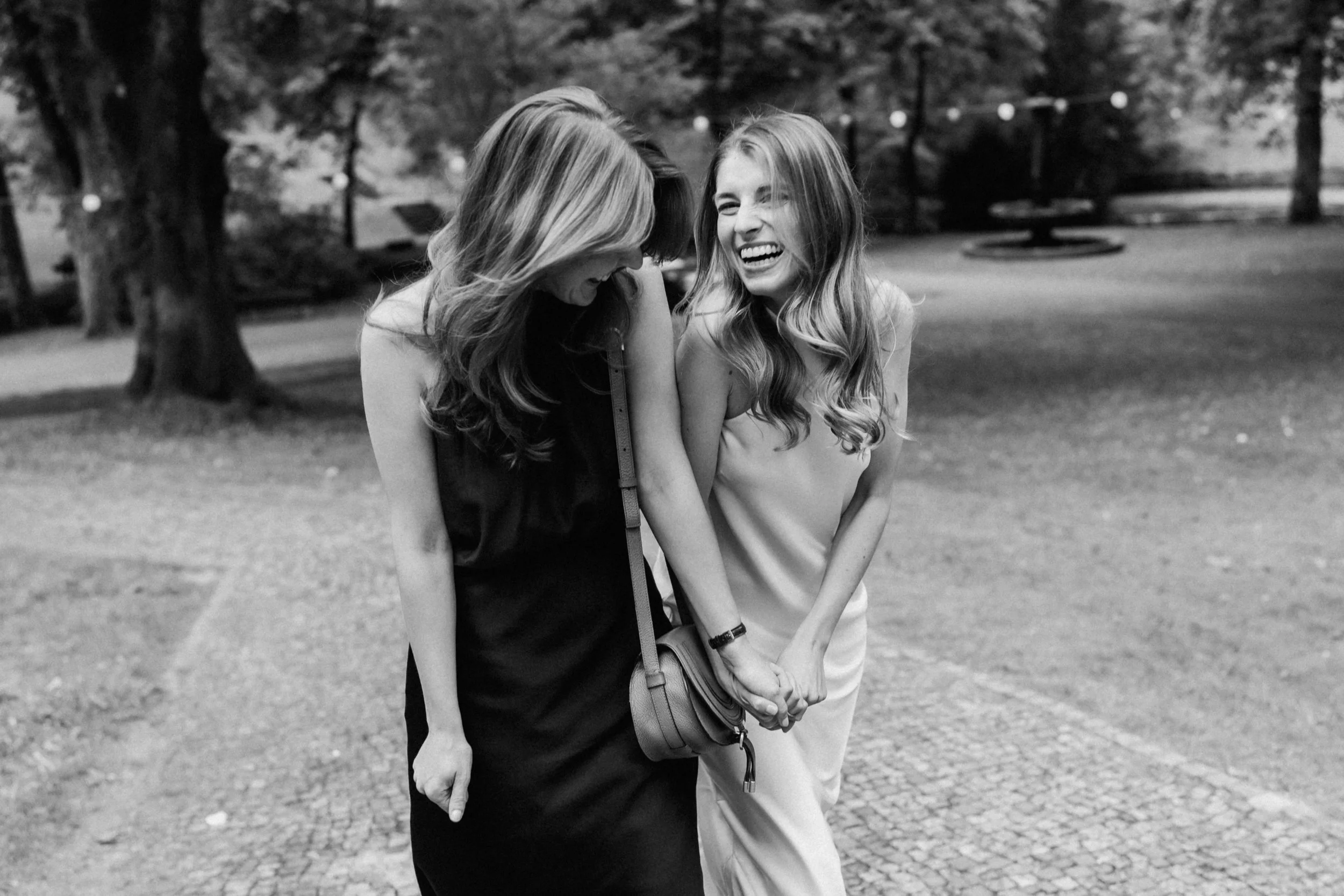 Two young women with long hair laughing and smiling while walking in a park on a summer day. One woman is wearing a black sleeveless top and the other a sleeveless top with light-colored pants. They are holding hands and appear to be enjoying each ot