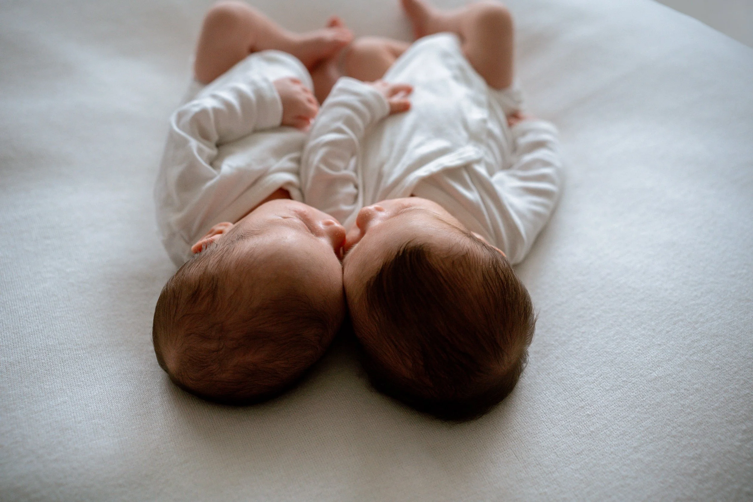 Two sleeping newborn babies lying on a white surface, side by side, dressed in white clothing.