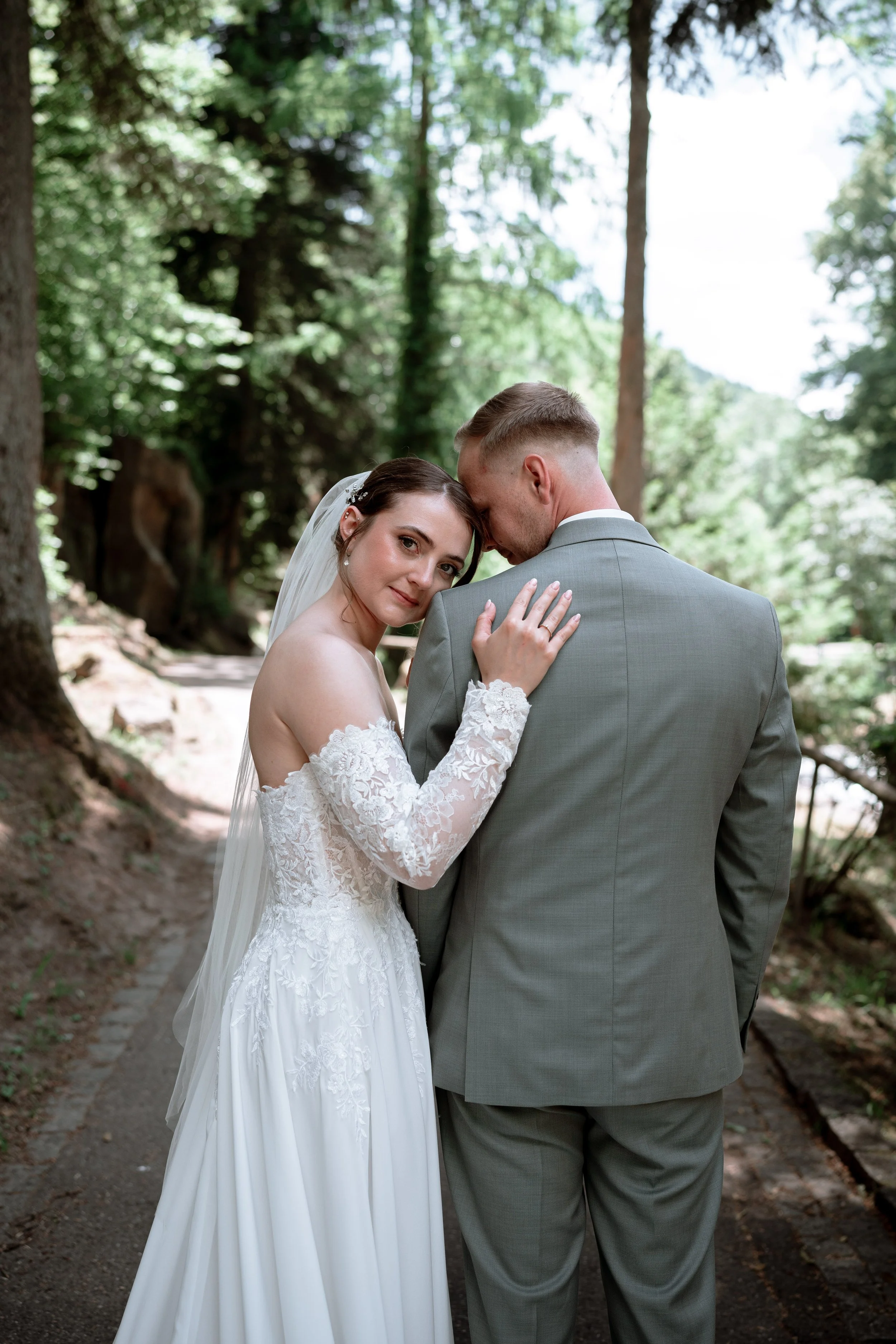 A bride and groom standing together on a forest trail, embracing with their foreheads touching. The bride is in a white lace wedding dress with long sleeves and a veil, looking at the camera. The groom is in a gray suit, facing away from the camera.