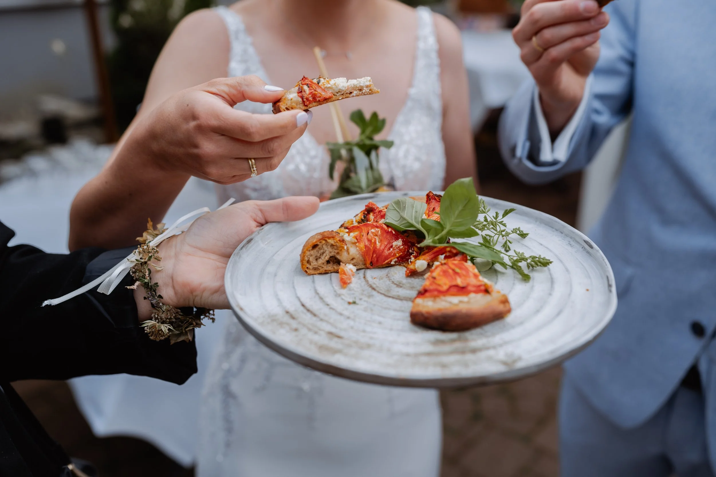 Person holding a half-eaten slice of pizza with visible toppings, serving pizza on a decorative white plate with herbs, at a social gathering.