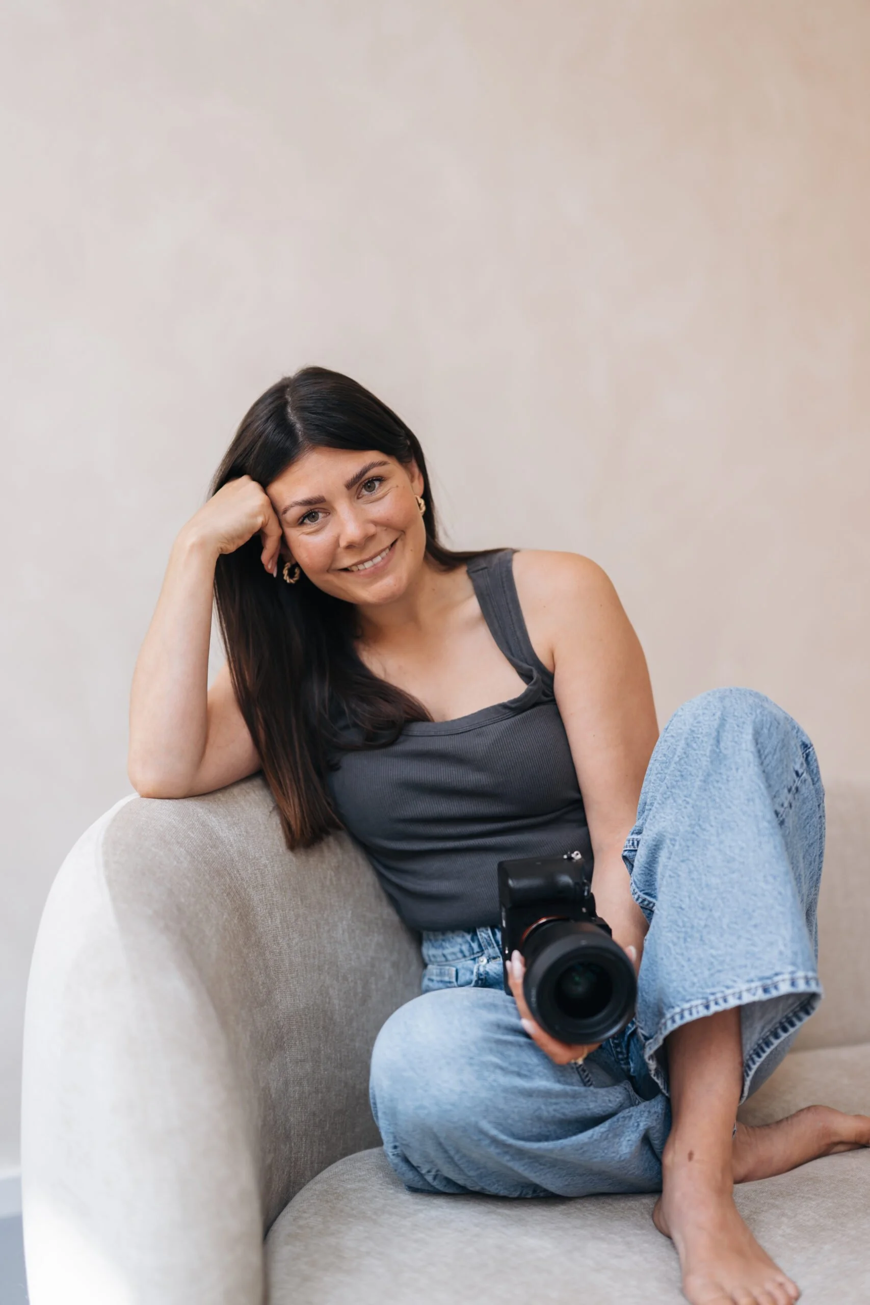 A woman with dark brown hair, wearing a gray tank top and blue jeans, sitting on a beige armchair. She is smiling, resting her head on her hand, holding a camera in her lap, with one leg crossed over the other and barefoot, against a plain white wall background.