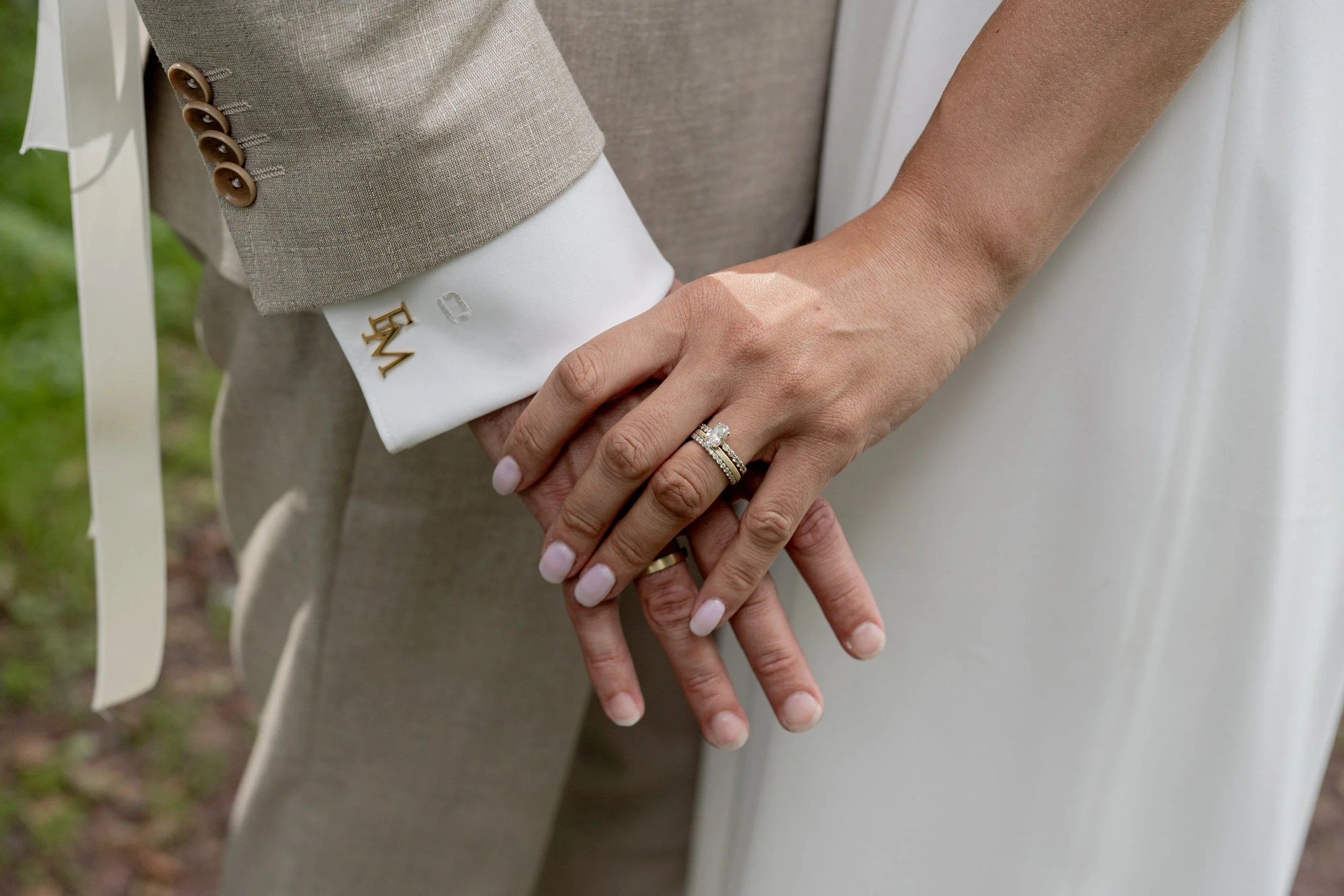 Close-up of a couple holding hands, with wedding rings visible on the woman's finger. The woman is wearing a white dress, and the man is wearing a beige suit with brown buttons on the sleeve. The background is blurred, suggesting an outdoor setting.