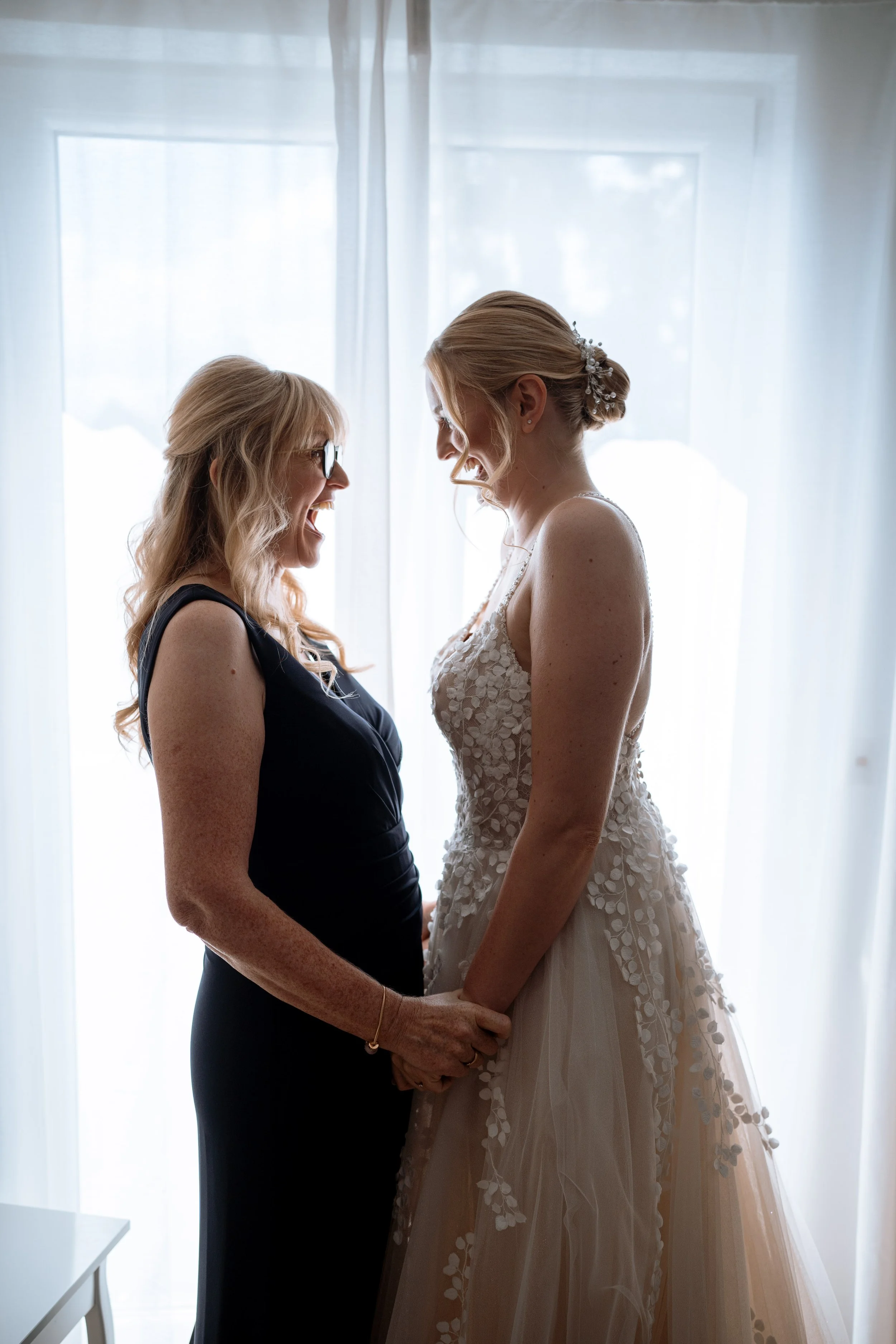 A woman in a black dress and eyeglasses and a woman in a wedding dress holding hands and smiling at each other in front of a window with sheer curtains.