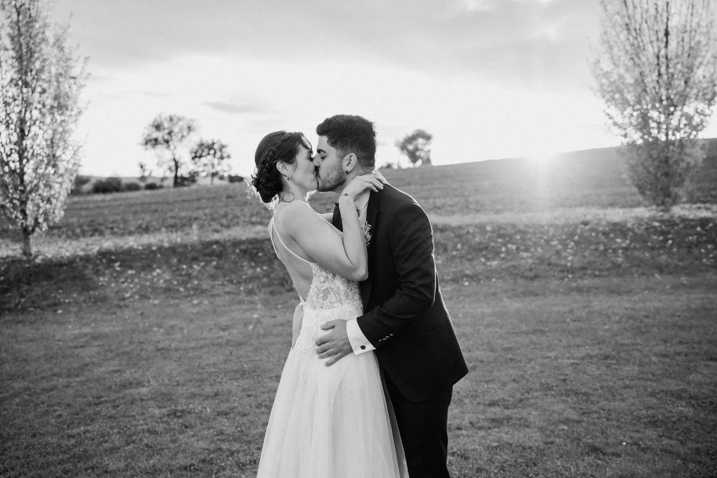 A couple in wedding attire sharing a kiss outdoors in a grassy field with trees and a cloudy sky in the background.