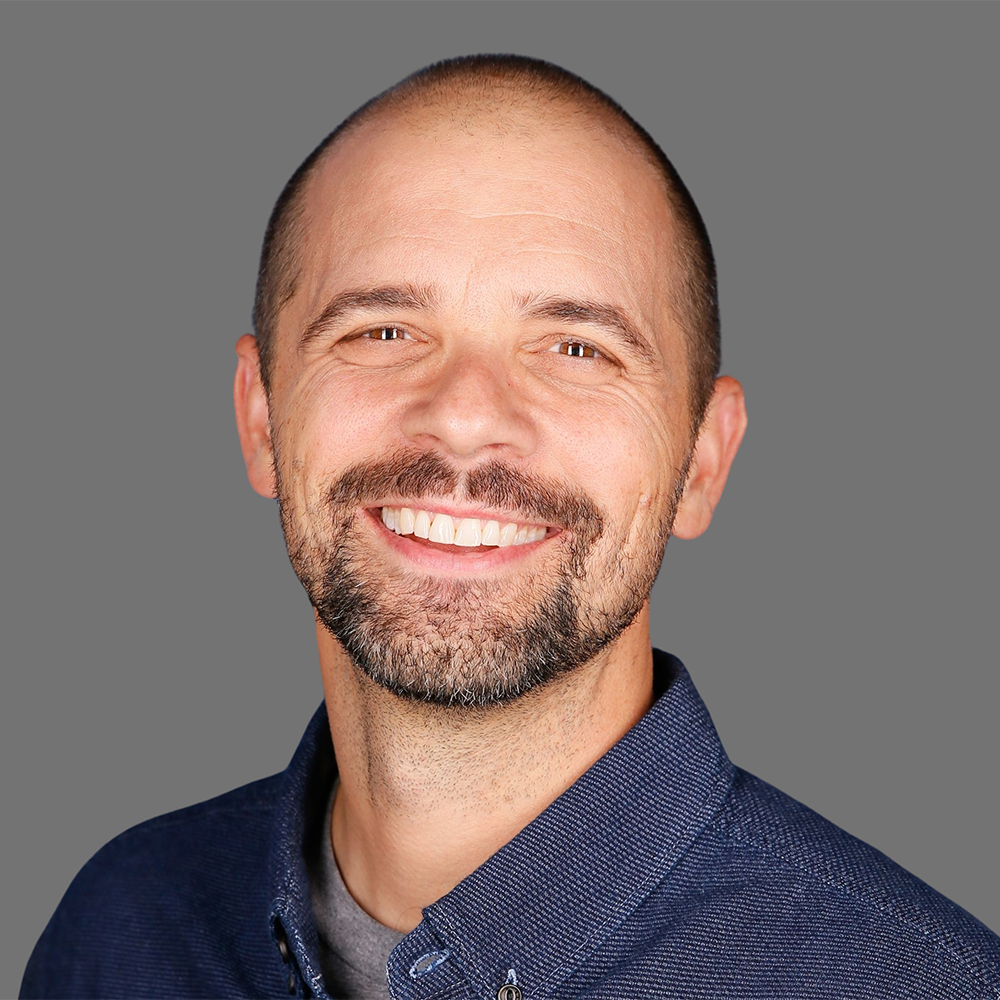 A smiling man with a beard and shaved head, wearing a dark blue collared shirt, against a gray background.
