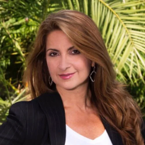 Woman with long brown hair wearing a black blazer and white top, standing outdoors with green tropical plants in the background.