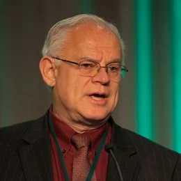 Older man with glasses and gray hair speaking, wearing a suit and tie, in front of a teal and brown background.