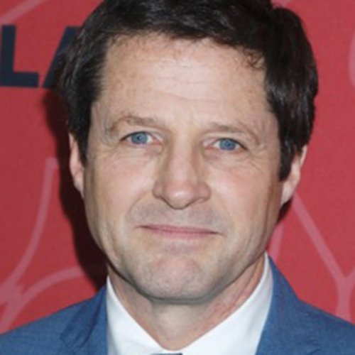 A close-up of a middle-aged man with dark hair, blue eyes, wearing a blue suit and white shirt, smiling in front of a red background.