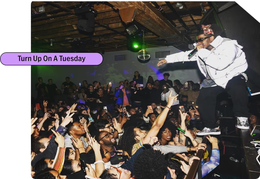 A performer on stage in a nightclub engaging with a crowd of people, with many reaching towards him. The nightclub is dimly lit with some green lights and a purple text overlay says, "Turn Up On A Tuesday."