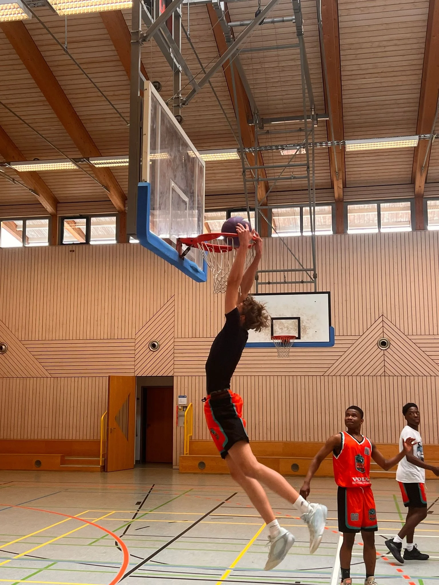 Ein Basketballspieler springt in einem Hallenbasketballfeld, um einen Korb zu erzielen, während andere Spieler in der Nähe stehen und zuschauen.