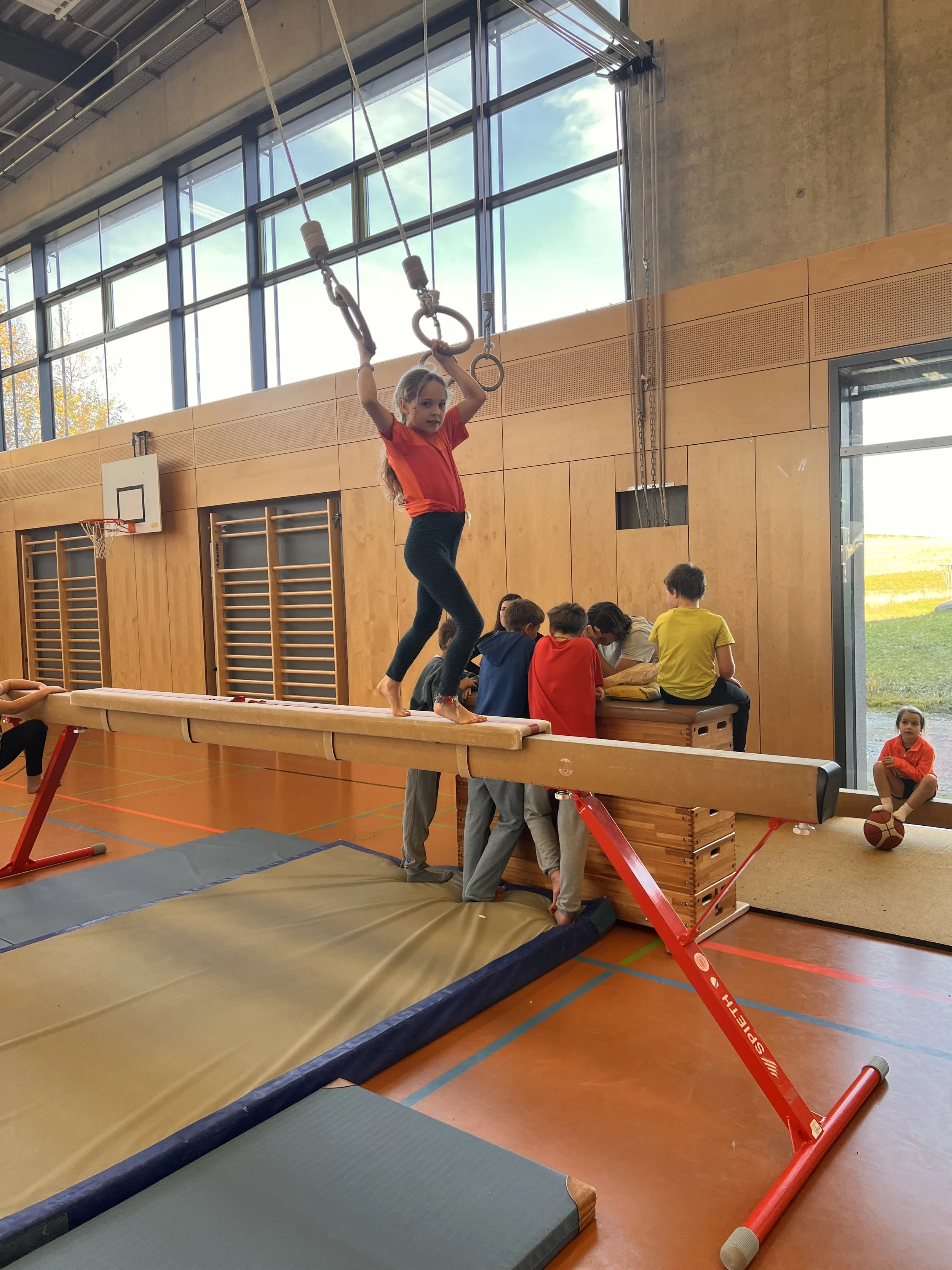 Ein Mädchen trainiert auf einer Turnbahn im Turnraum der Turnhalle, während andere Kinder im Hintergrund auf einer Bank sitzen.