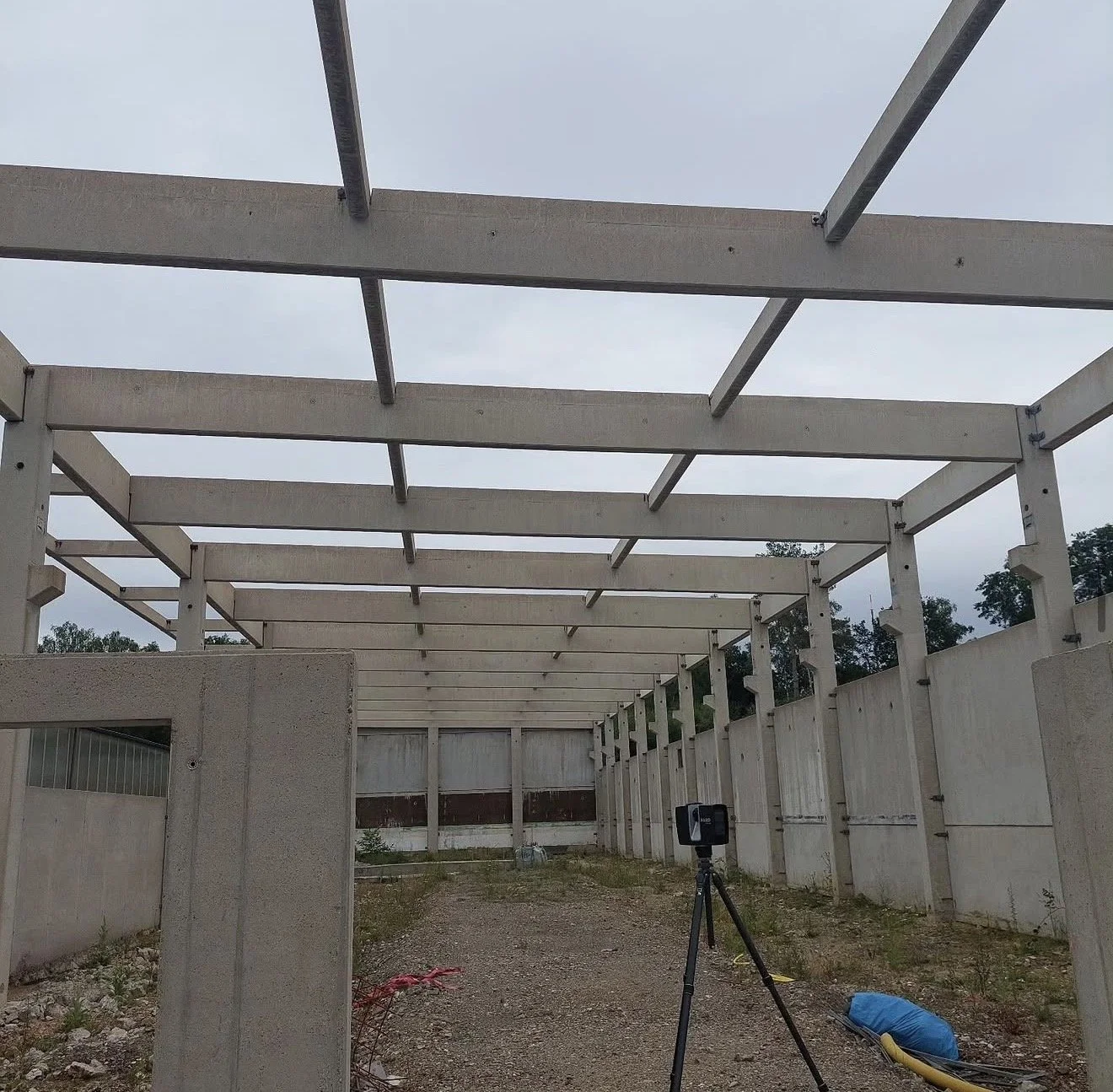 Baustelle mit weißem Konstruktionsrahmen, Bauarbeiten im Gange, Baugerät und Materialien auf dem Boden, überwachende Kamera auf Stativ, mit Bäumen im Hintergrund.