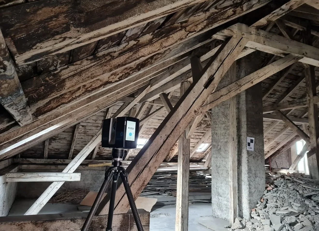 Innenaufnahme eines Dachbodens mit Holzbalken, Baustoffen und einem Laser-Scanner auf einem Stativ.
