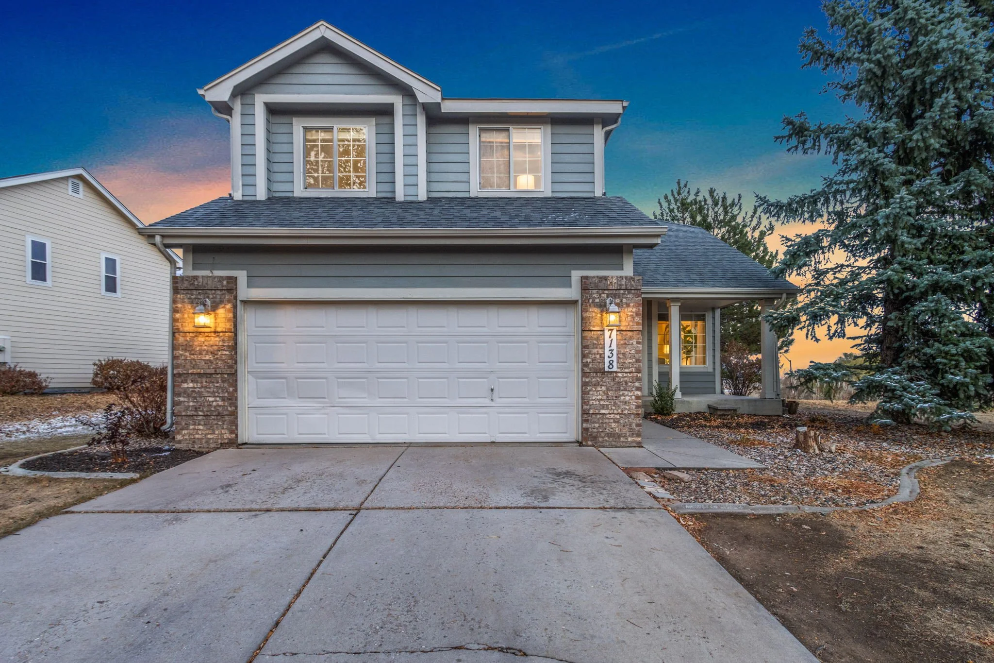 Front view of a two-story house with a white garage door, brick accents, and gray siding, illuminated by outdoor lights at dusk, with trees and a colorful sky in the background.