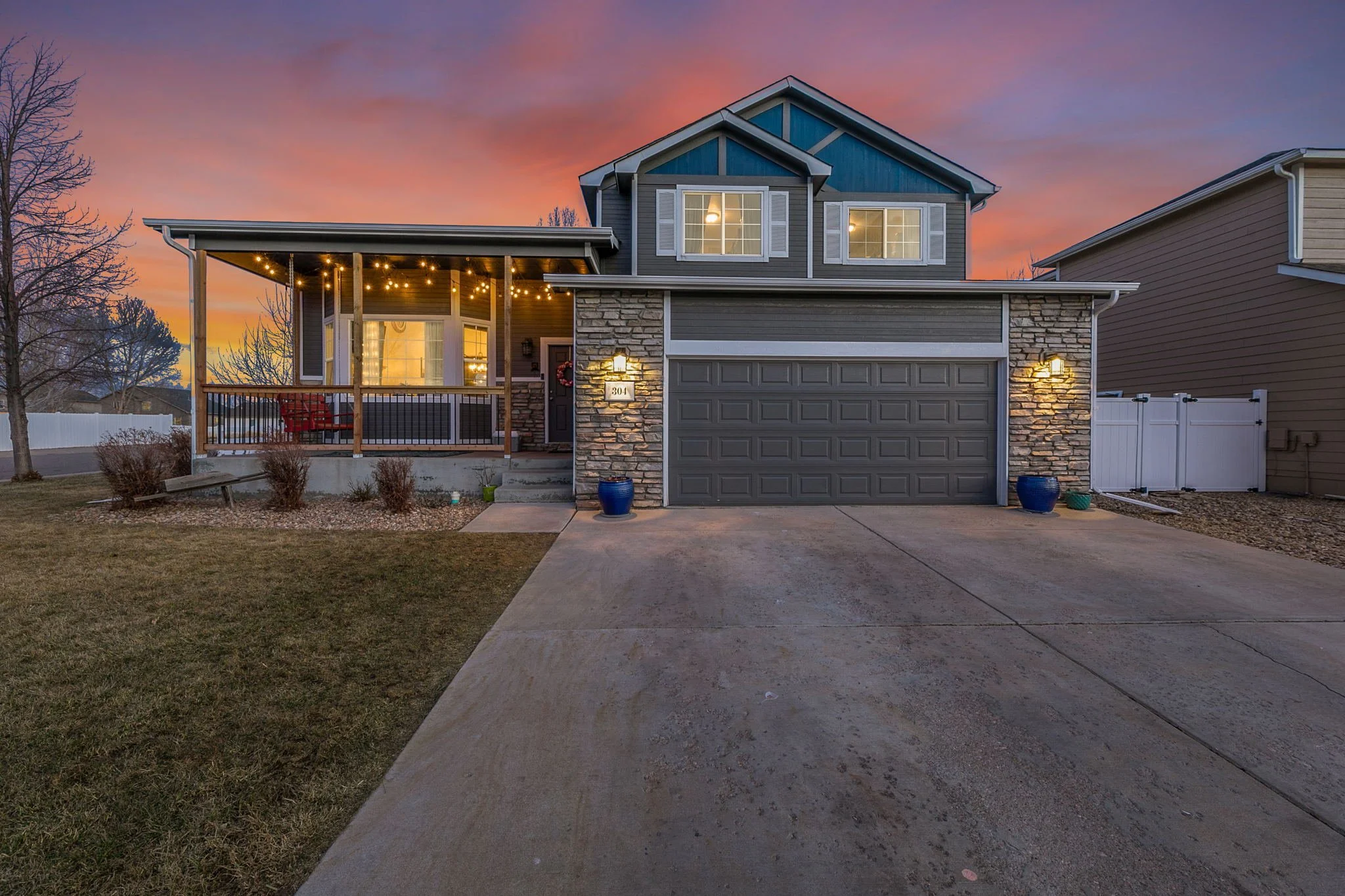 Front view of a two-story gray house with a garage, porch with string lights, and a sunset sky in the background.