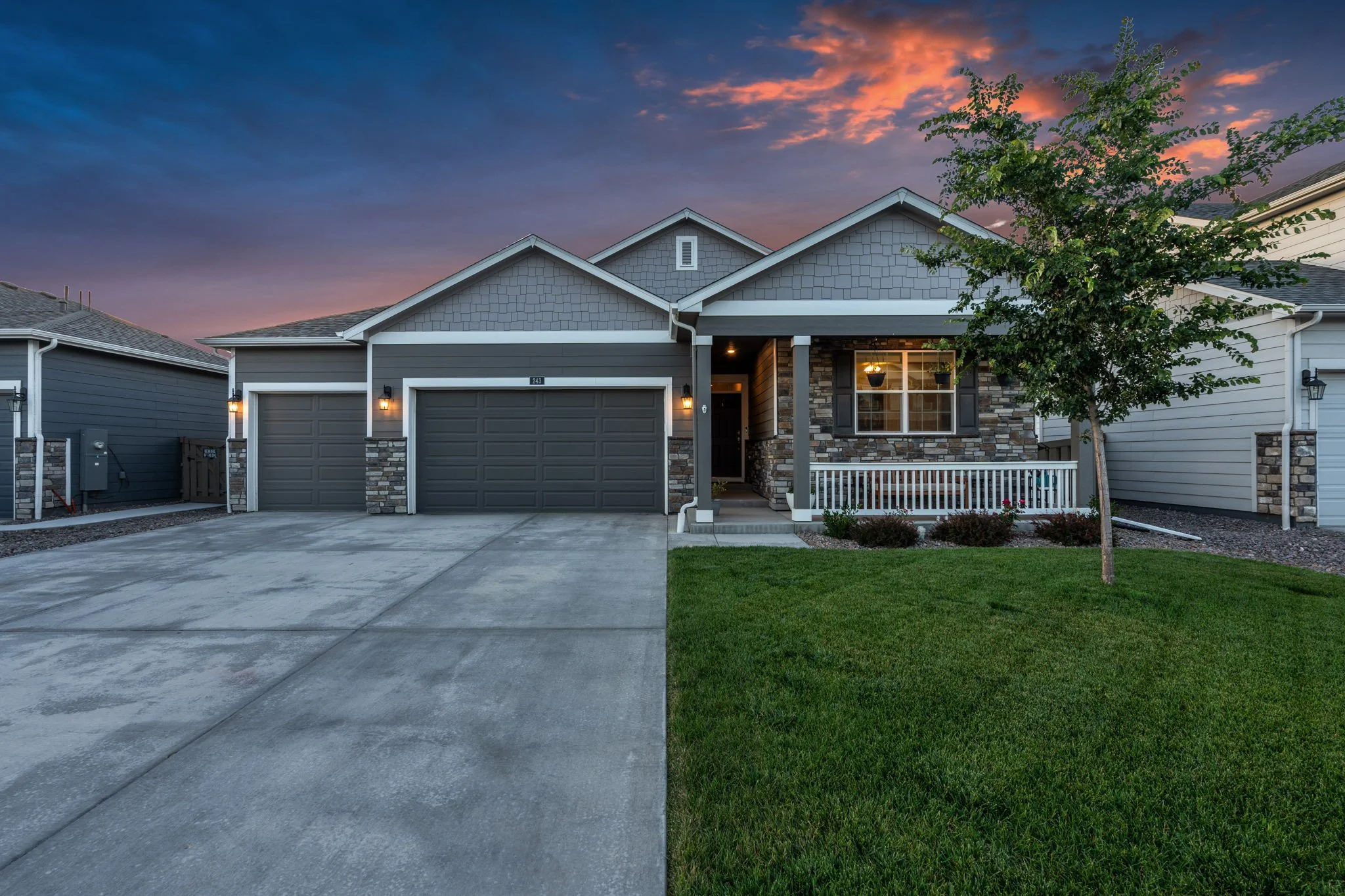 A modern suburban house with a dark gray garage door, stone and gray siding exterior, a front porch with a white railing, a small tree with green leaves, a well-maintained lawn, and a colorful sunset sky in the background.