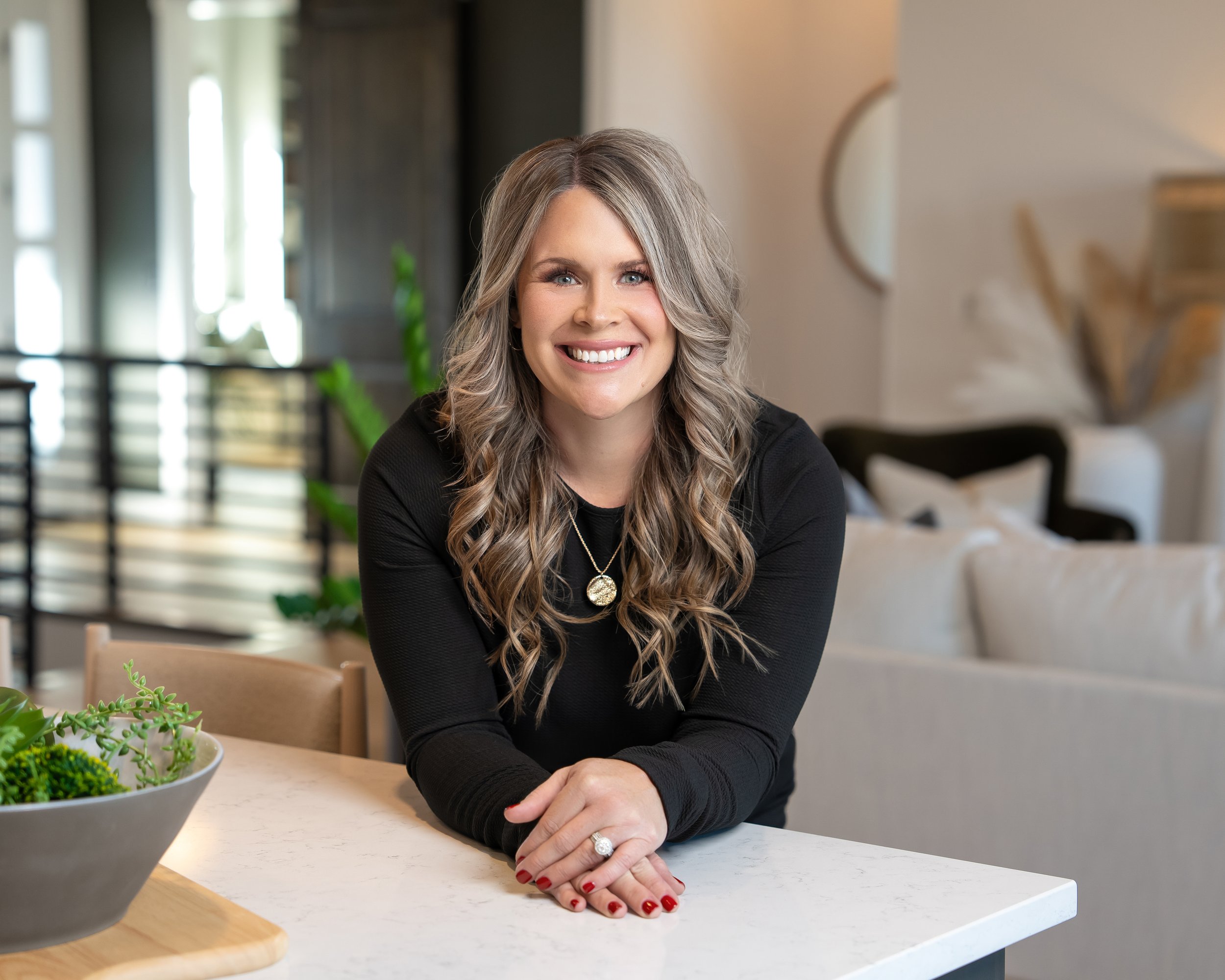 A smiling woman with long wavy gray hair sitting at a kitchen island with her hands clasped on the countertop. She is wearing a black top and a ring, with red nail polish. There is a bowl of succulents on the counter in front of her, and the background shows a modern kitchen with plants and a window.
