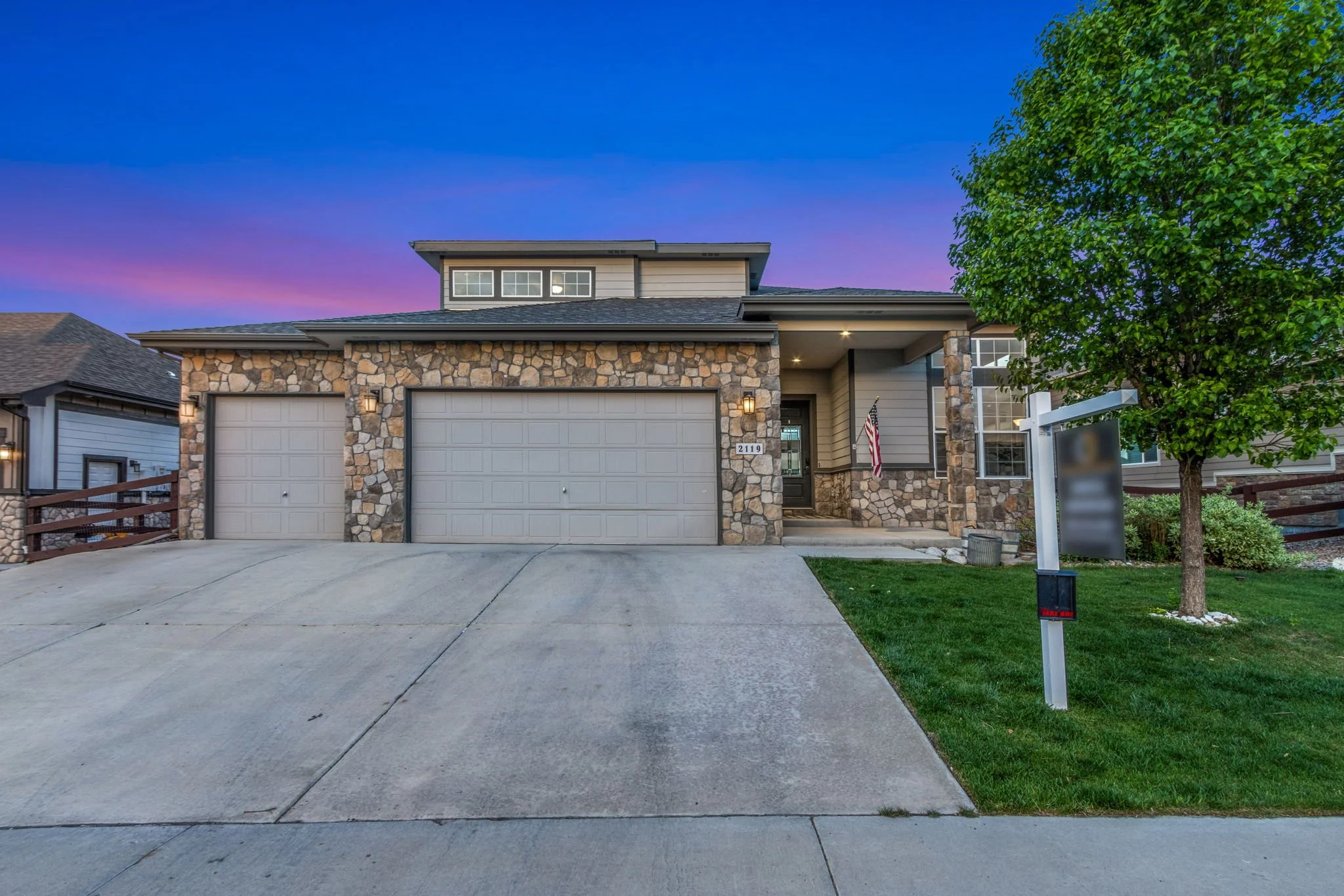 Front view of a two-story house with stone and siding exterior, a garage, a small front porch, trees, and a blemished driveway at dusk with a purple and blue sky.