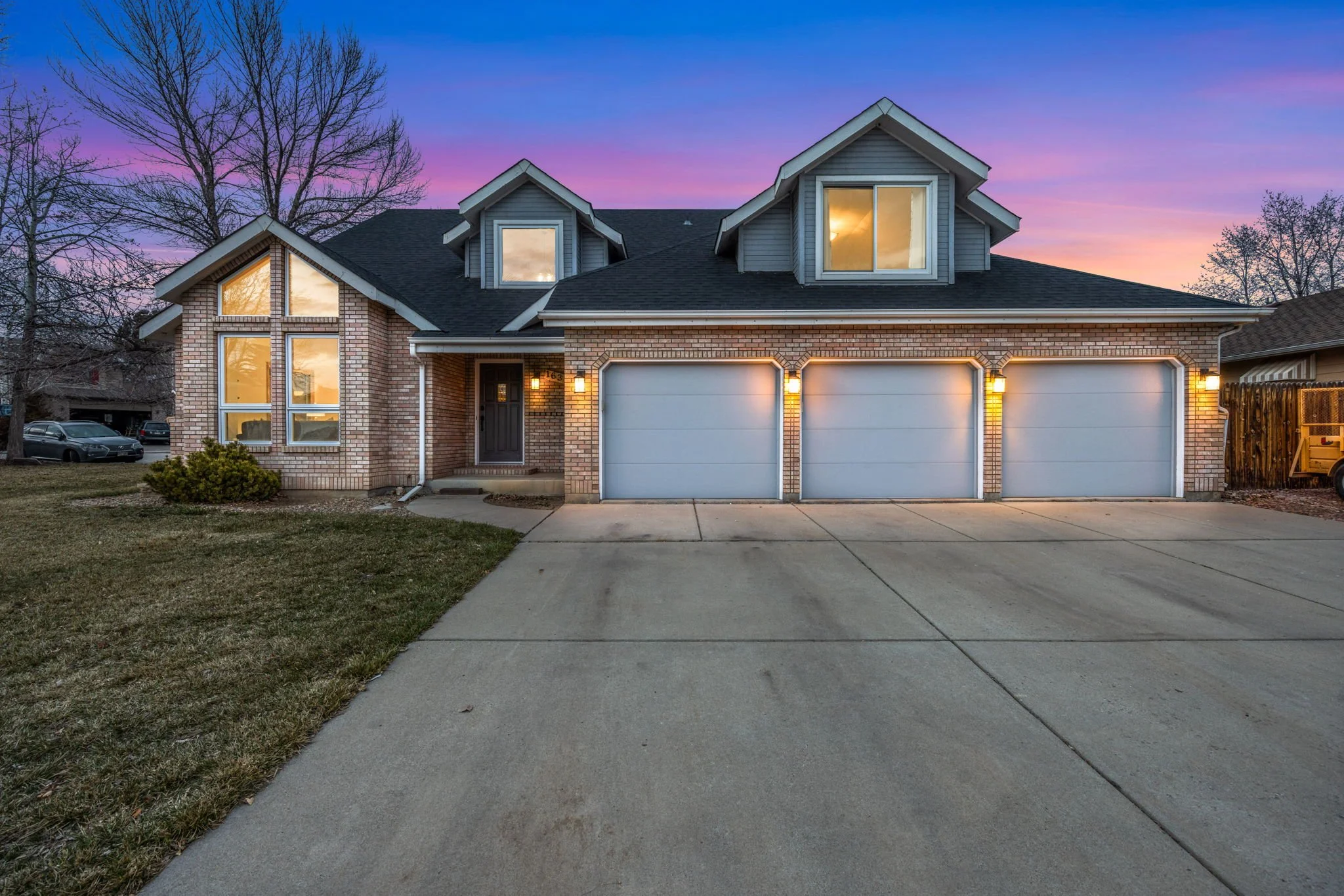 Front view of a suburban house at dusk with brick and light blue siding, three dormer windows, illuminated porch light, and closed garage doors, with trees and parked cars in the background.