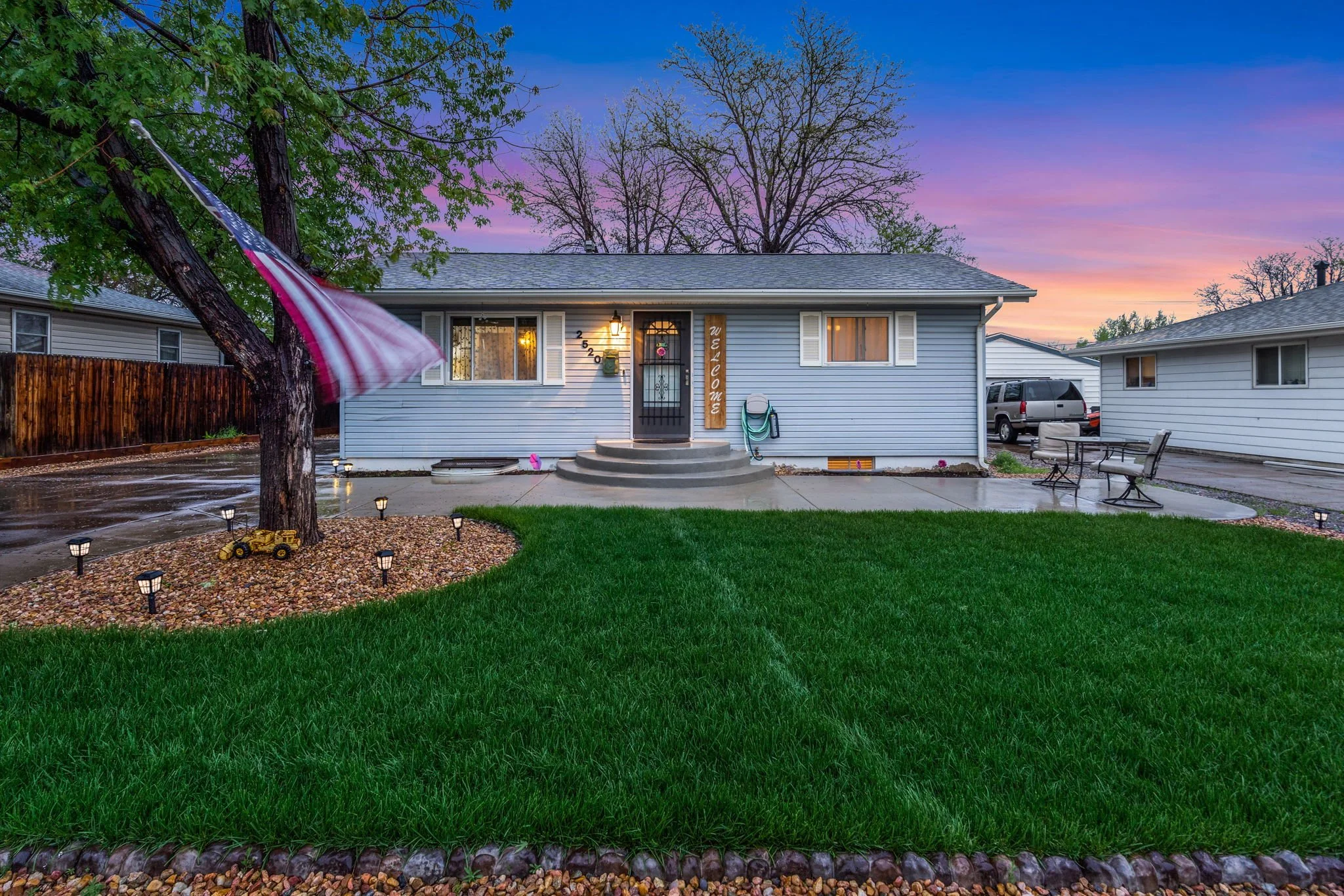 Front yard of a house with a lawn, a tree with an American flag, a pebble garden, small lights, and a porch with steps at sunset.