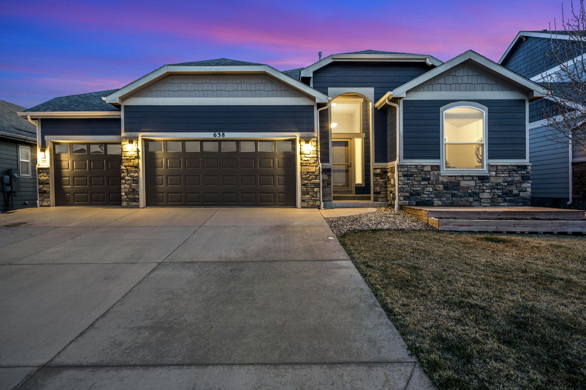 A two-story house with blue siding, stone accents, and a double garage door, illuminated by exterior lighting during dusk with a colorful sky.