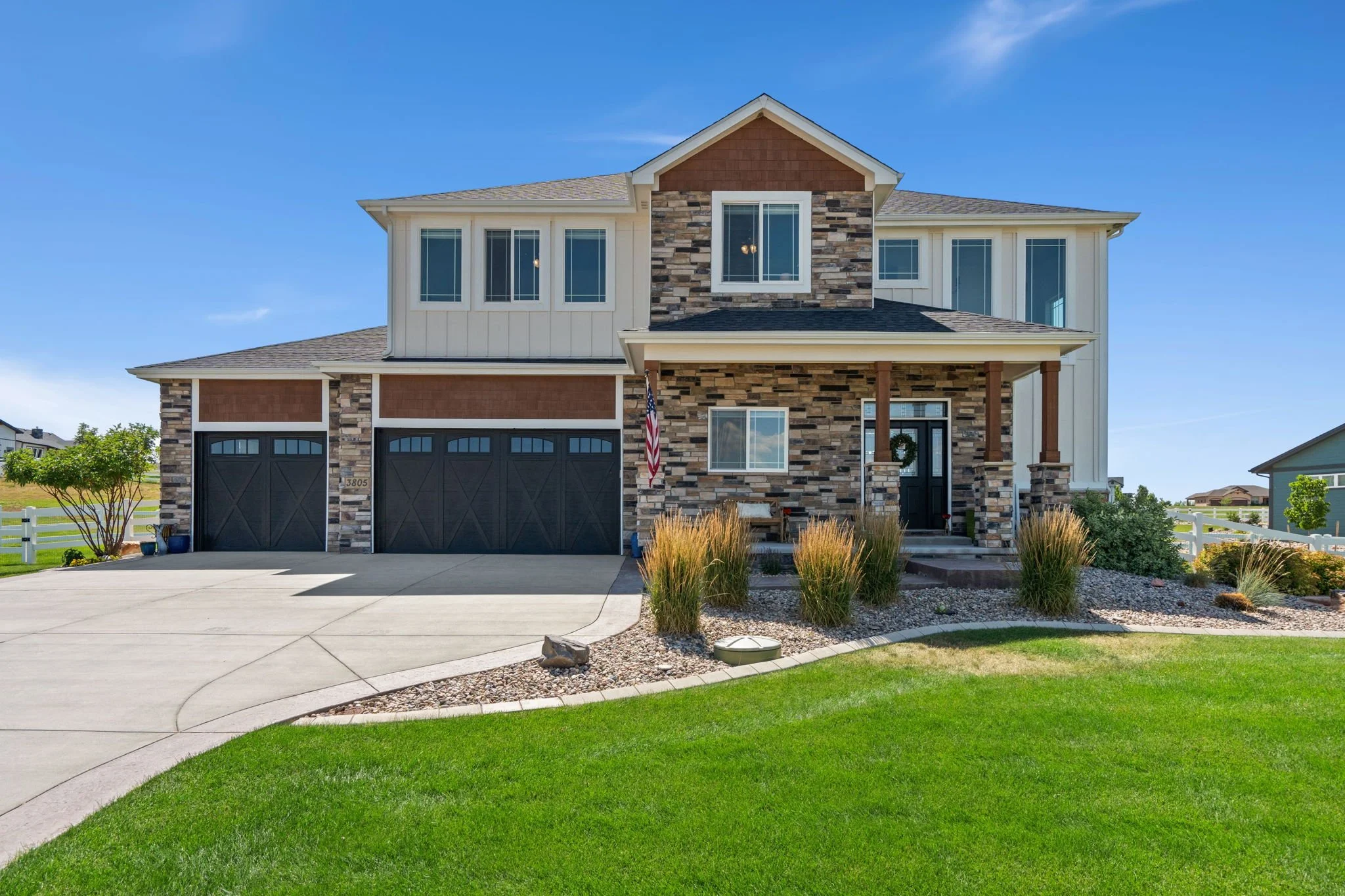 Front view of a two-story house with a stone and siding exterior, black garage doors, front porch, and green lawn.