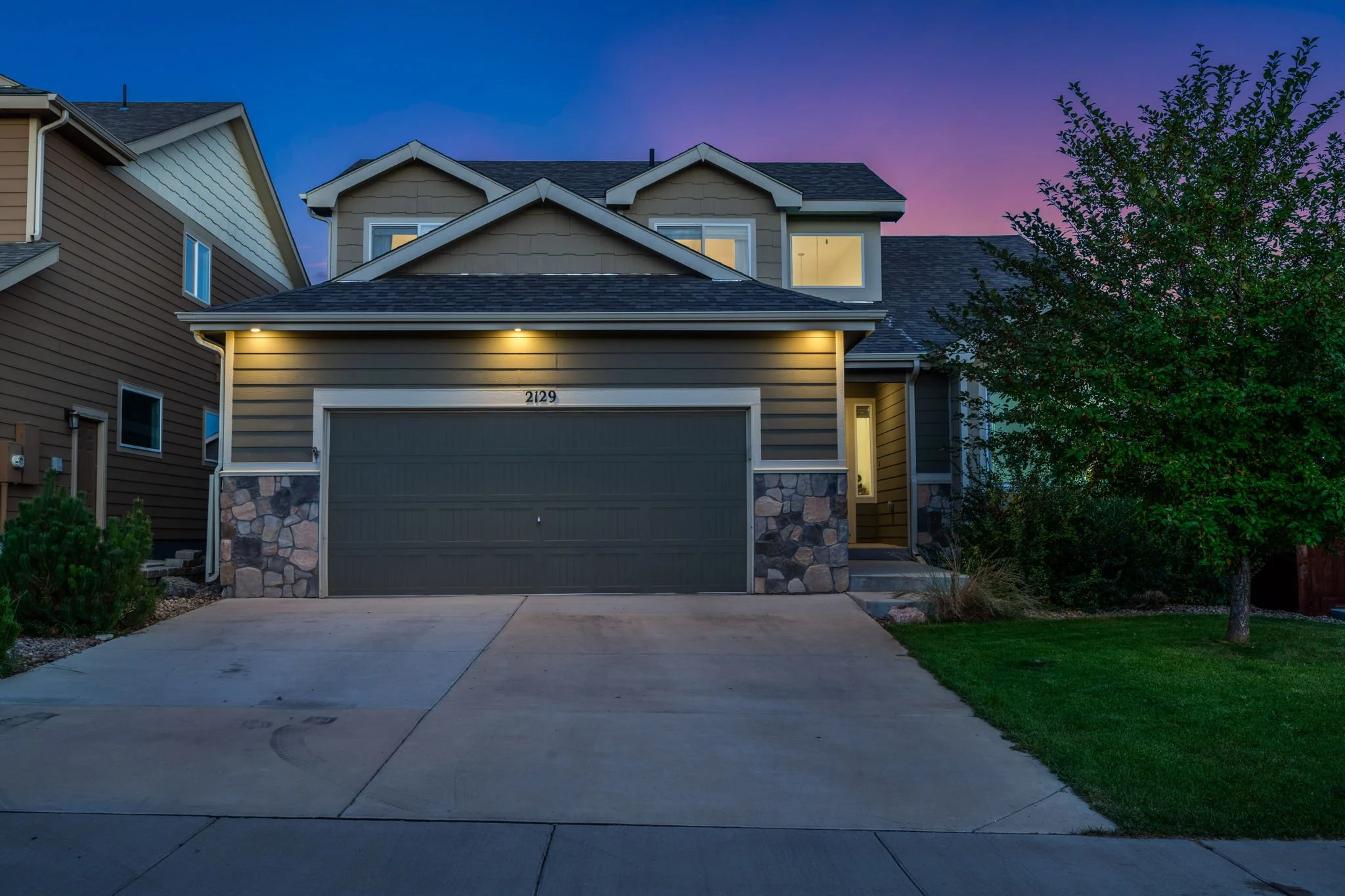 A modern house with a two-car garage, lit porch lights, and a small front yard with grass and a tree, during twilight evening.