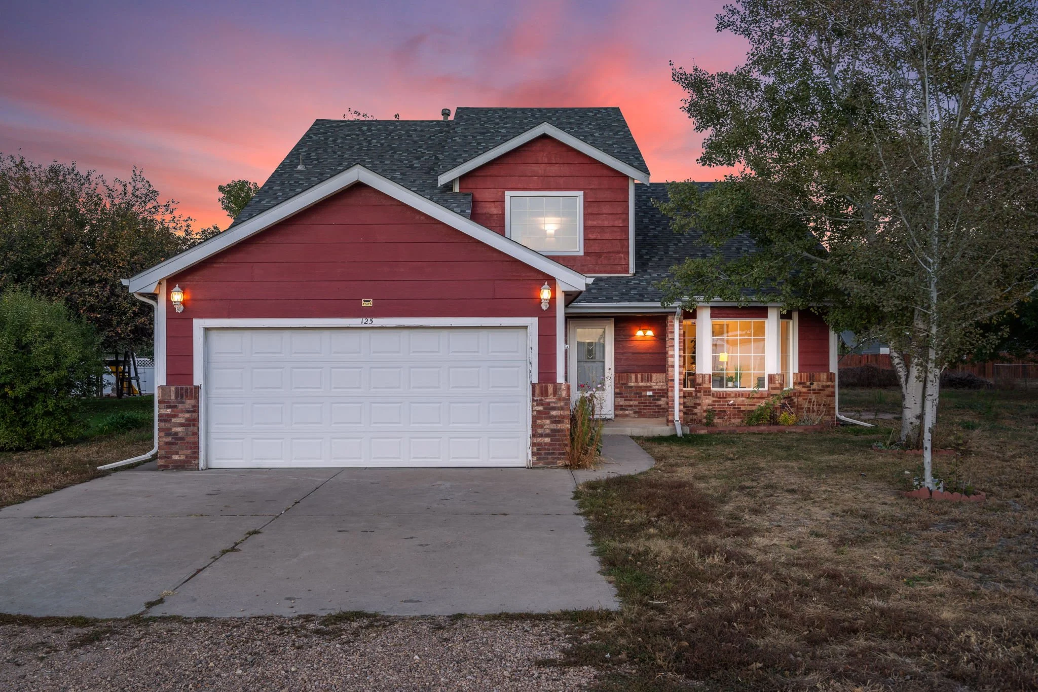 A two-story red house with a garage and an illuminated porch, surrounded by trees at sunset, with a cloudy pink and purple sky.