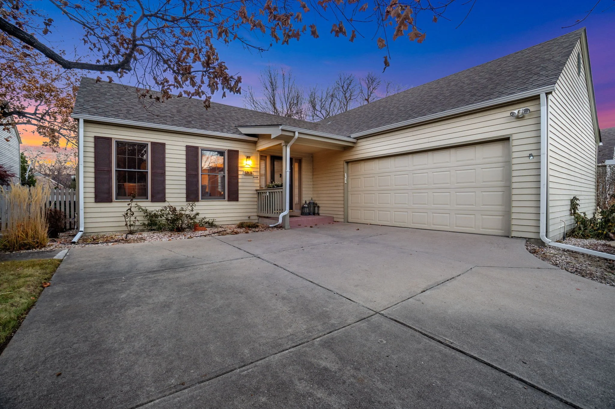 A beige house with a two-car garage, front porch, brown shutters, and a lit exterior light at dusk, surrounded by trees with bare branches and a paved driveway.