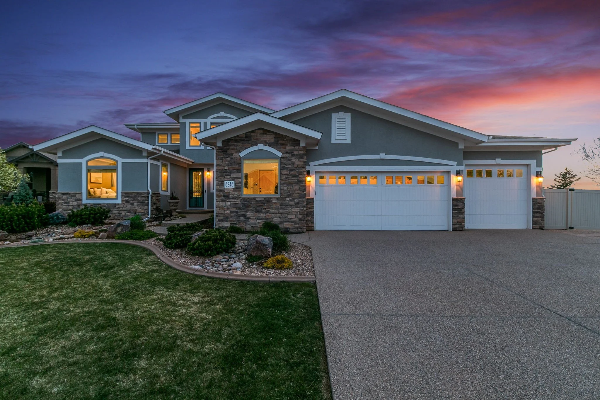 A modern two-story house with a stone and blue stucco exterior, illuminated windows, and a white garage door, set against a colorful evening sky with a purple and orange sunset.