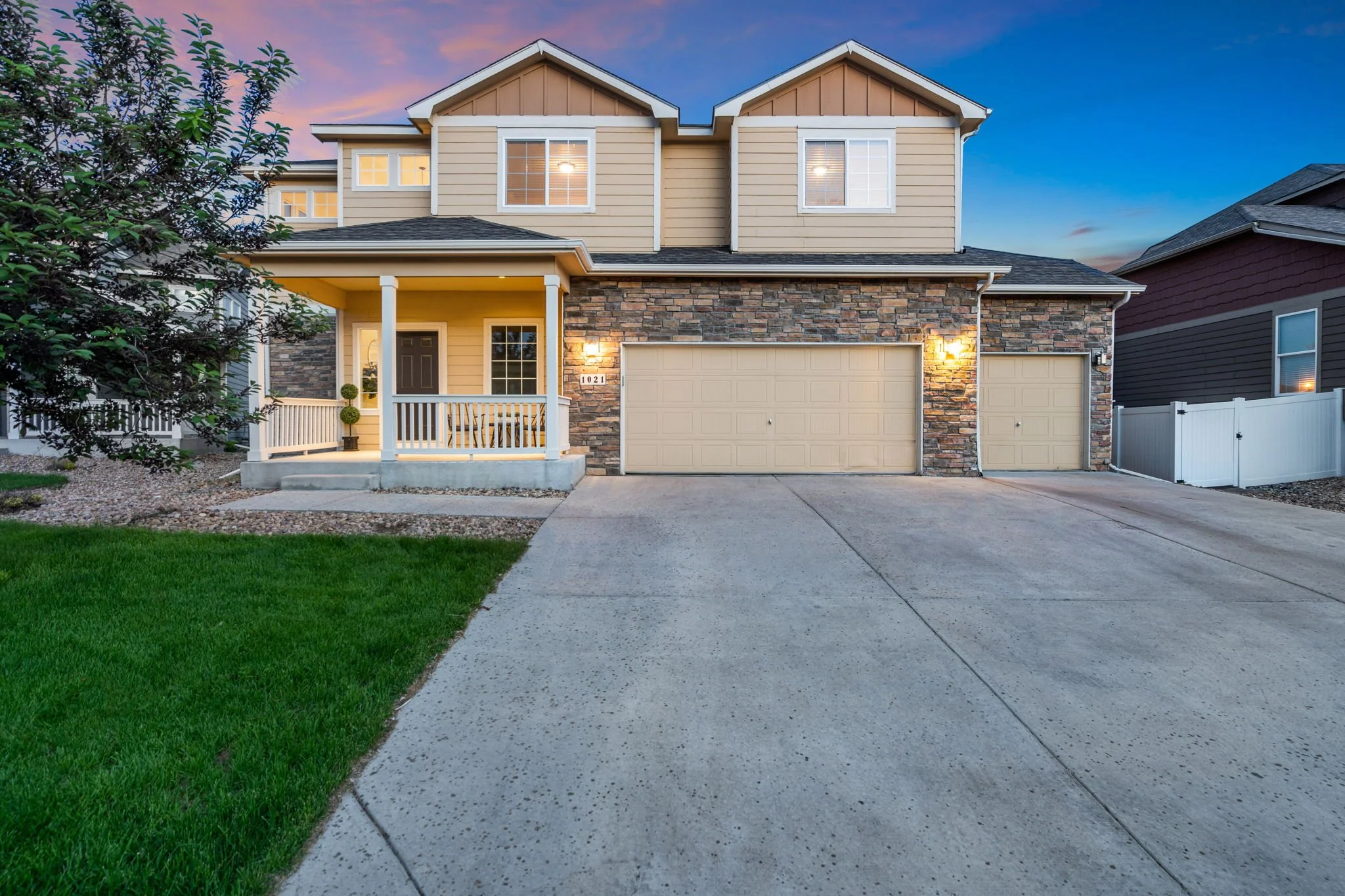 A two-story house with beige siding, stone accents, and a dark shingled roof. The front yard has green grass, and there is a concrete driveway. The house has a porch with white railings and a black front door. Windows are lit from inside, and the sky