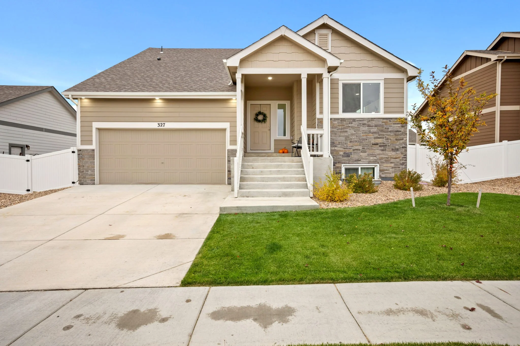 A beige two-story house with a front porch, stone accents on the lower part, white trim, and a gabled roof. The house has a cream-colored garage door and steps leading to the front door that is decorated with a wreath. There are pumpkins on the porch