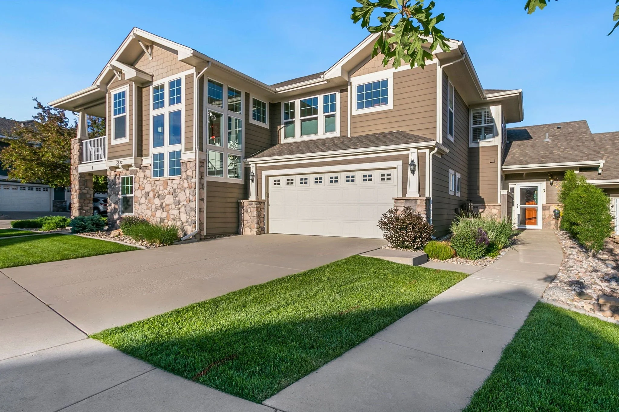 A modern two-story house with a brown exterior, multiple large windows, a white garage door, and a well-maintained lawn with landscaped plants and a concrete driveway.