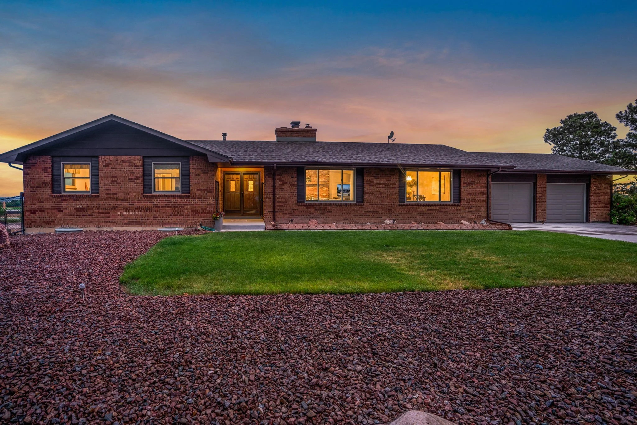 Rear view of a brick house at sunset with illuminated interior, two garage doors, four windows, a front door, and landscaped yard in the foreground.