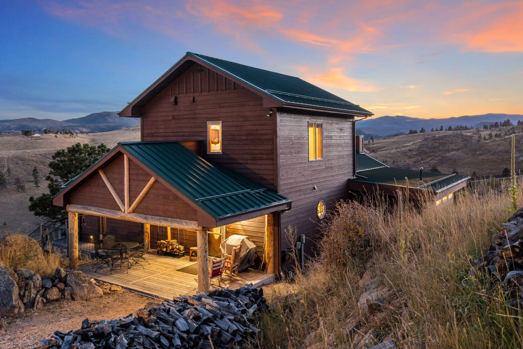 A modern wooden house with a metal roof, situated on a hillside at sunset, with mountains in the background. The house has a lit interior and an outdoor patio area with furniture.