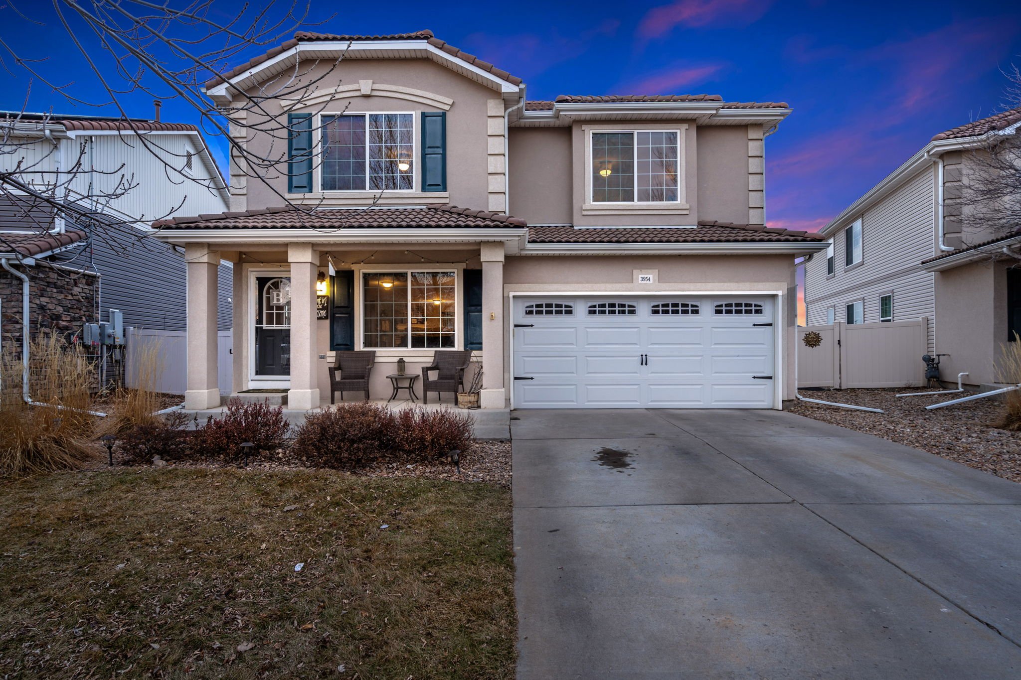 A two-story suburban house with a beige exterior, dark green shutters, a white garage door, and a small front porch with chairs, during dusk with a colorful sky in the background.