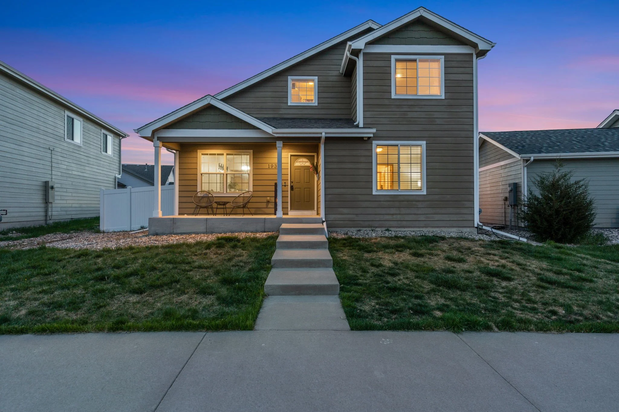 Front view of a two-story house with a porch, windows, and a covered entrance, during twilight or early evening with a colorful sky.