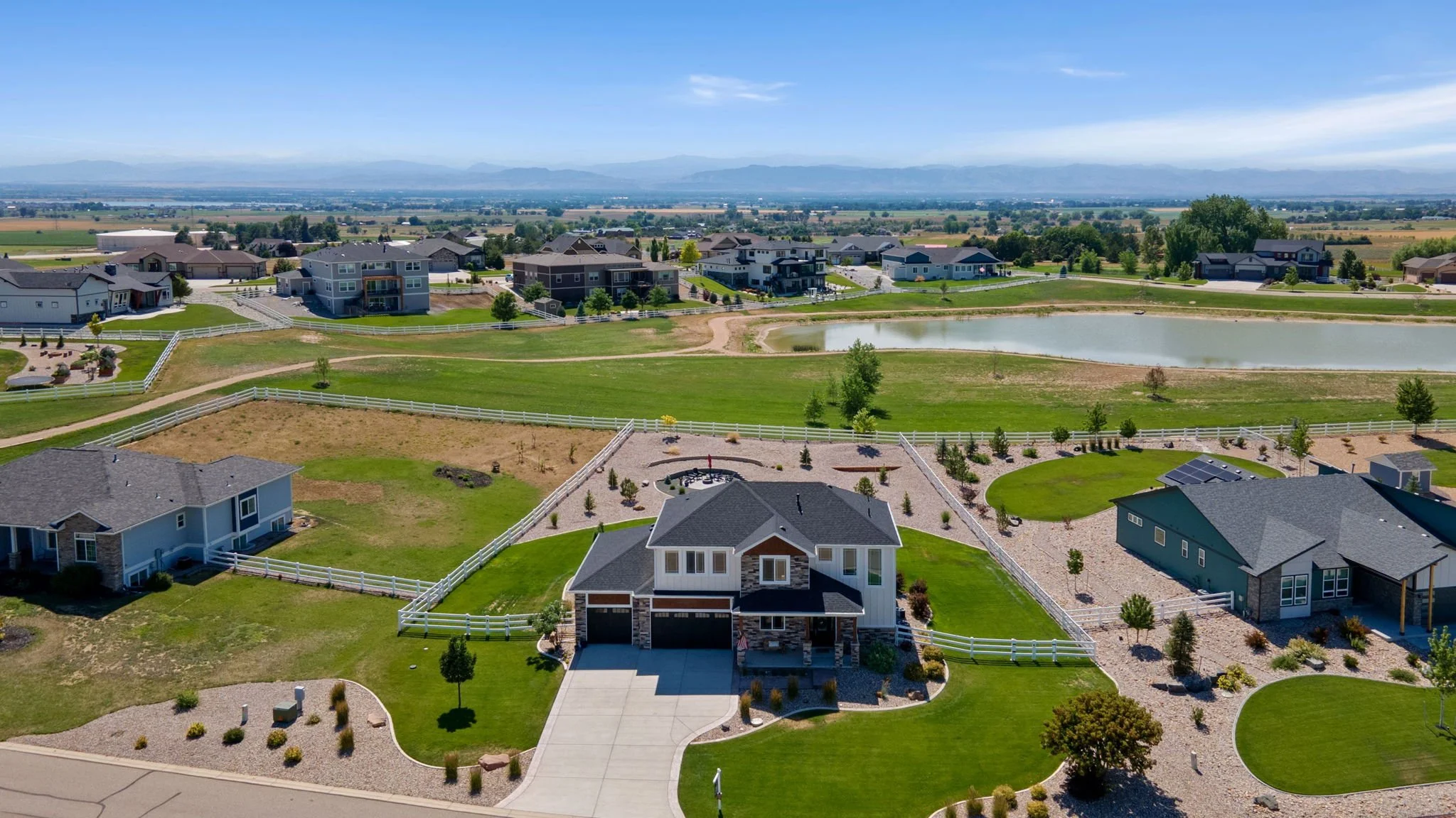 Aerial view of a suburban neighborhood with houses, green lawns, a pond, and distant mountains in the background under a clear blue sky.