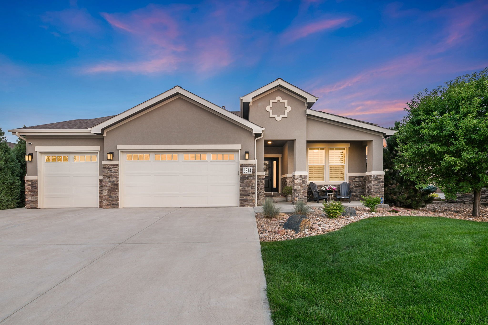 A modern single-family house with a three-car garage, stone accents, a small front porch with seating, and landscaped yard, under a colorful sunset sky.