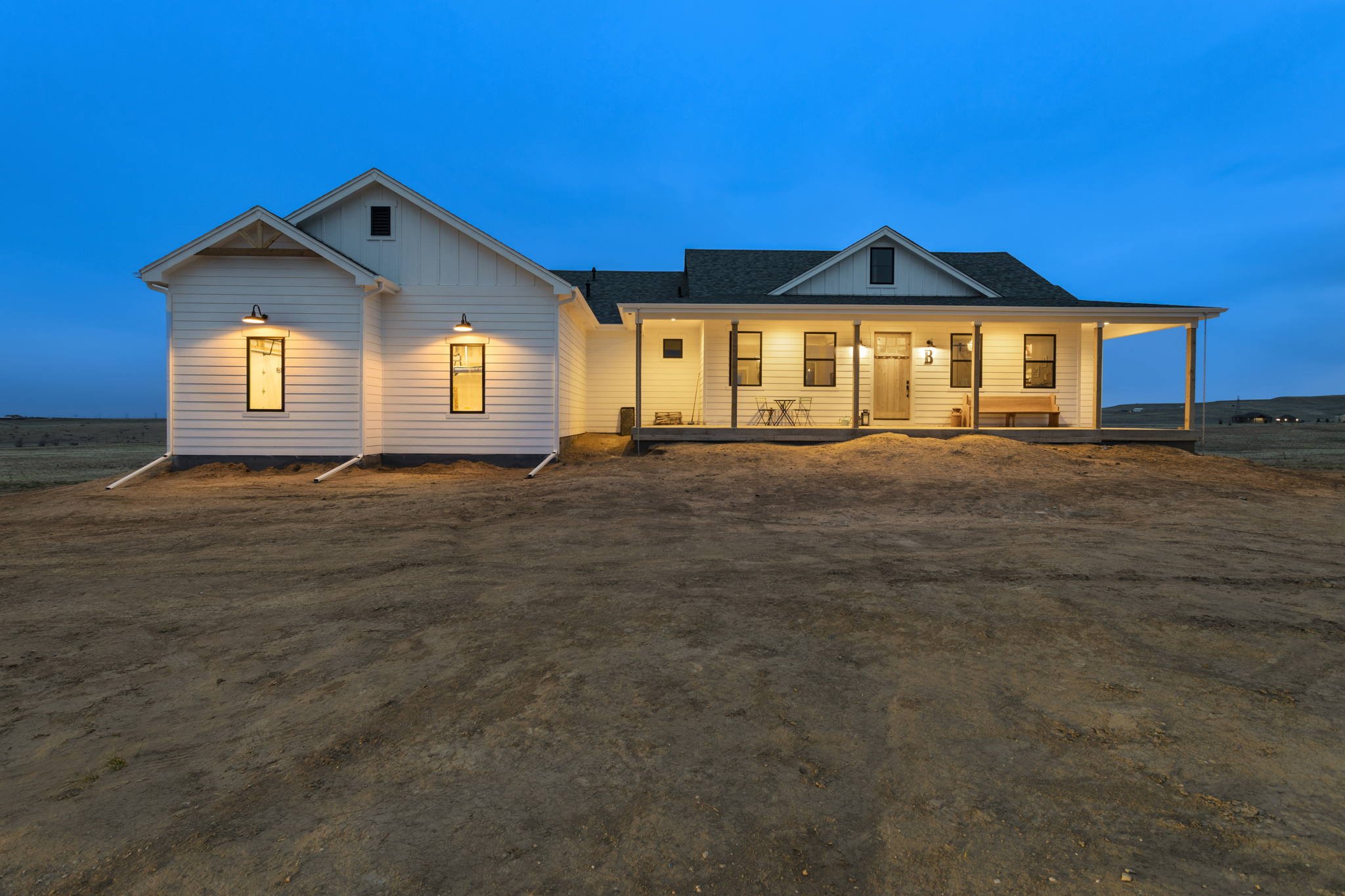 A white house with a covered porch and outdoor seating, illuminated with warm lights, situated in a flat, barren landscape during dusk.