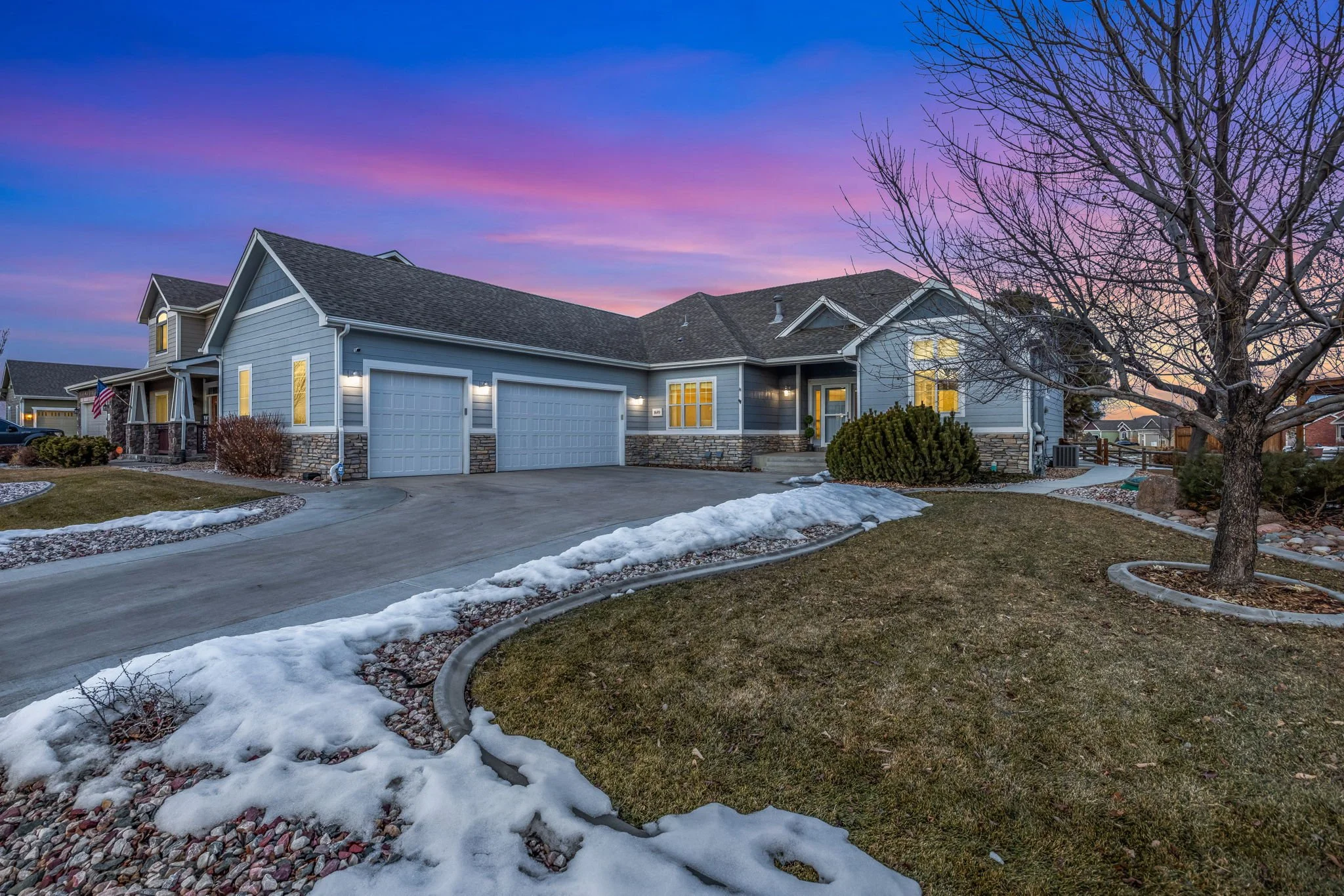 A suburban house during sunset with a colorful sky, snow patches on the lawn, leafless trees, and illuminated windows.