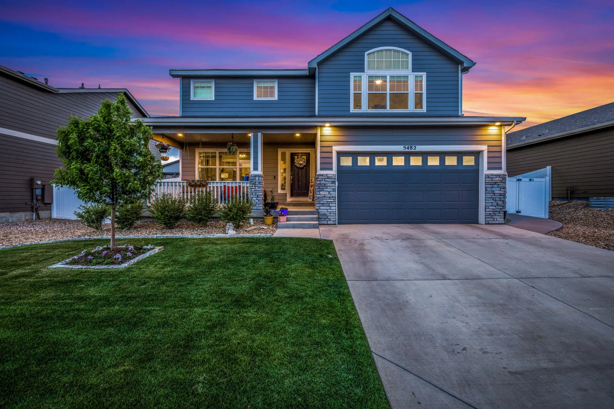 A two-story blue house with a well-kept front yard and a concrete driveway at sunset, with colorful sky in the background.