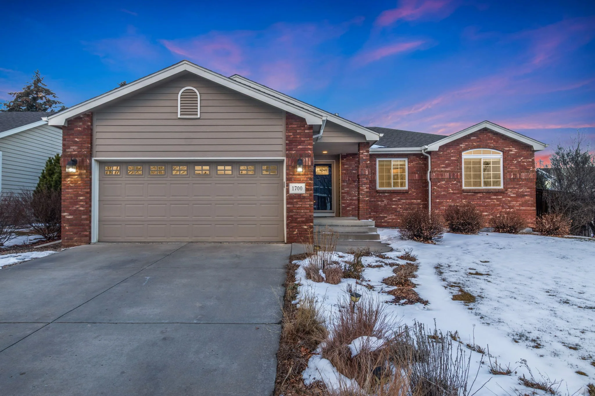 Front view of a house with a two-car garage, brick and siding exterior, and a small front yard partially covered with snow, under a colorful evening sky.