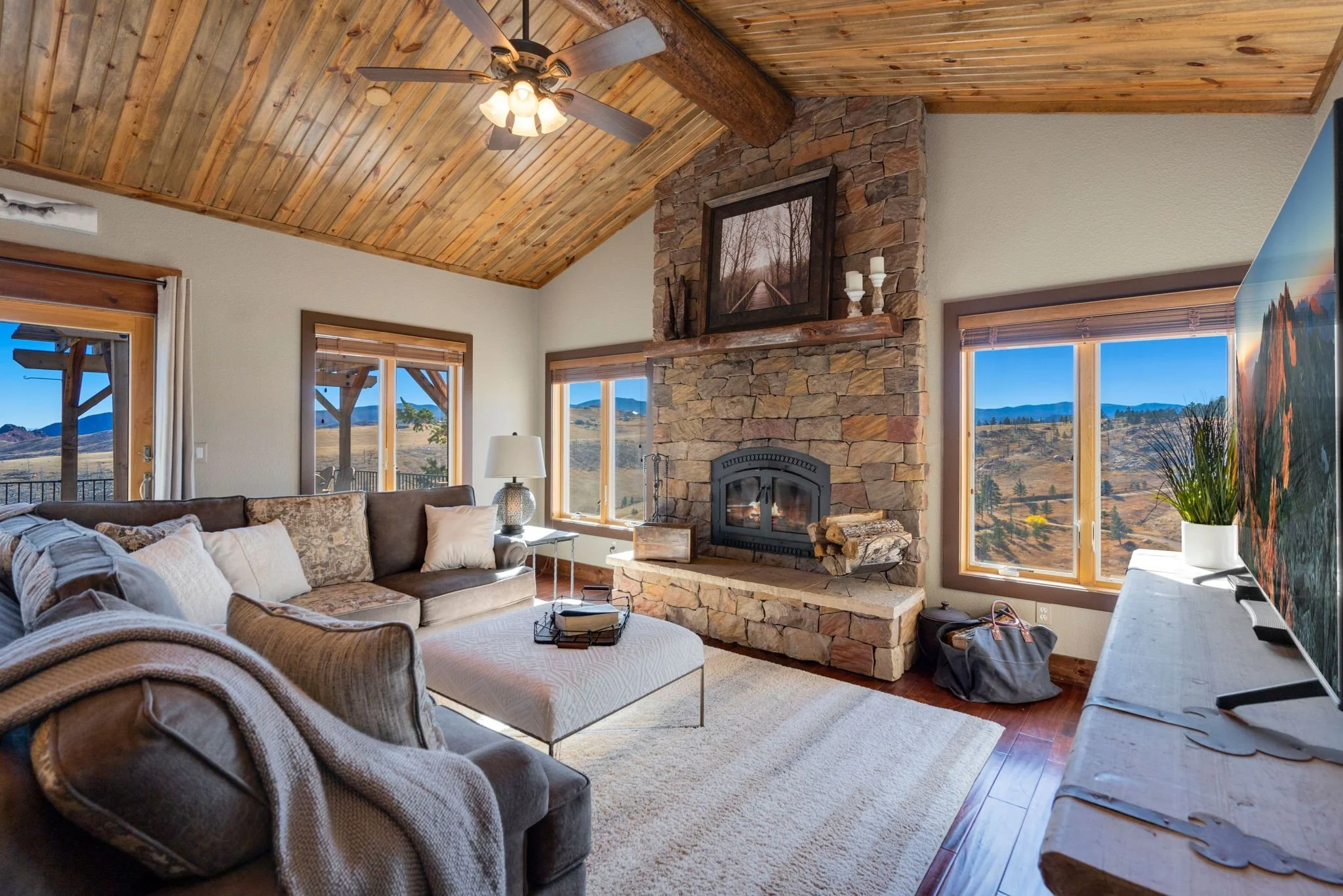 Living room with a vaulted wooden ceiling, a stone fireplace with a landscape painting above, large windows showing mountains and fields, and beige furniture with pillows and blankets.